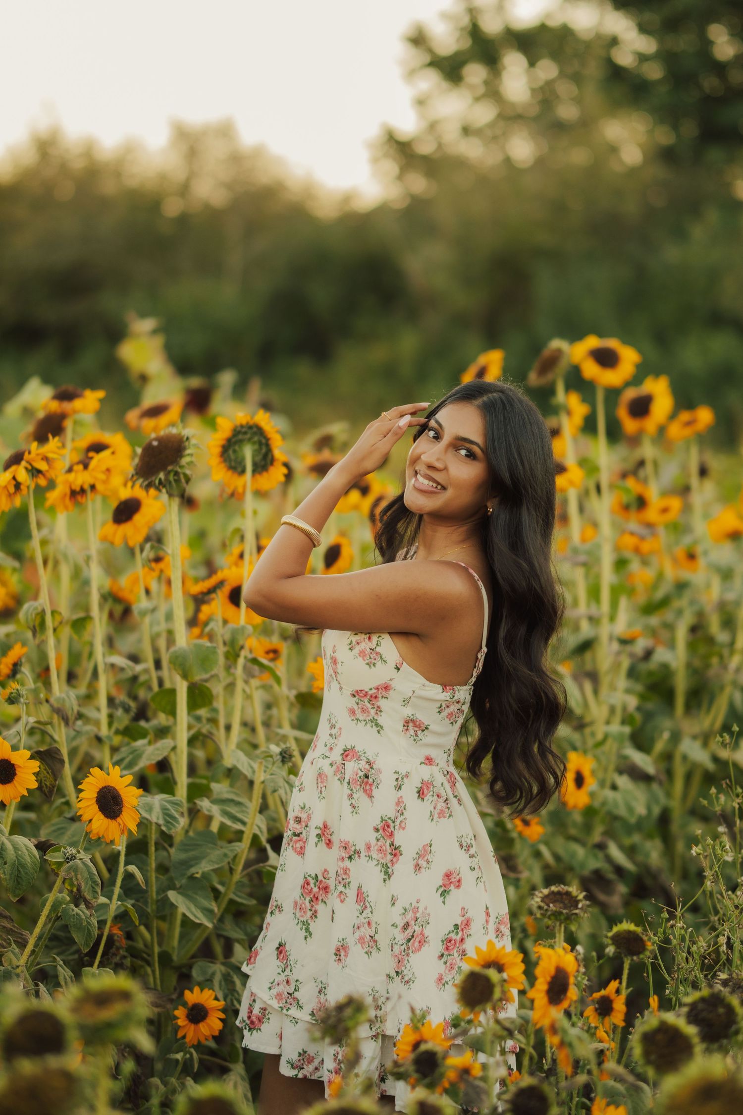 Woman with long dark hair in a floral sundress smiles and poses with her hand raised in a blooming sunflower field at golden hour.