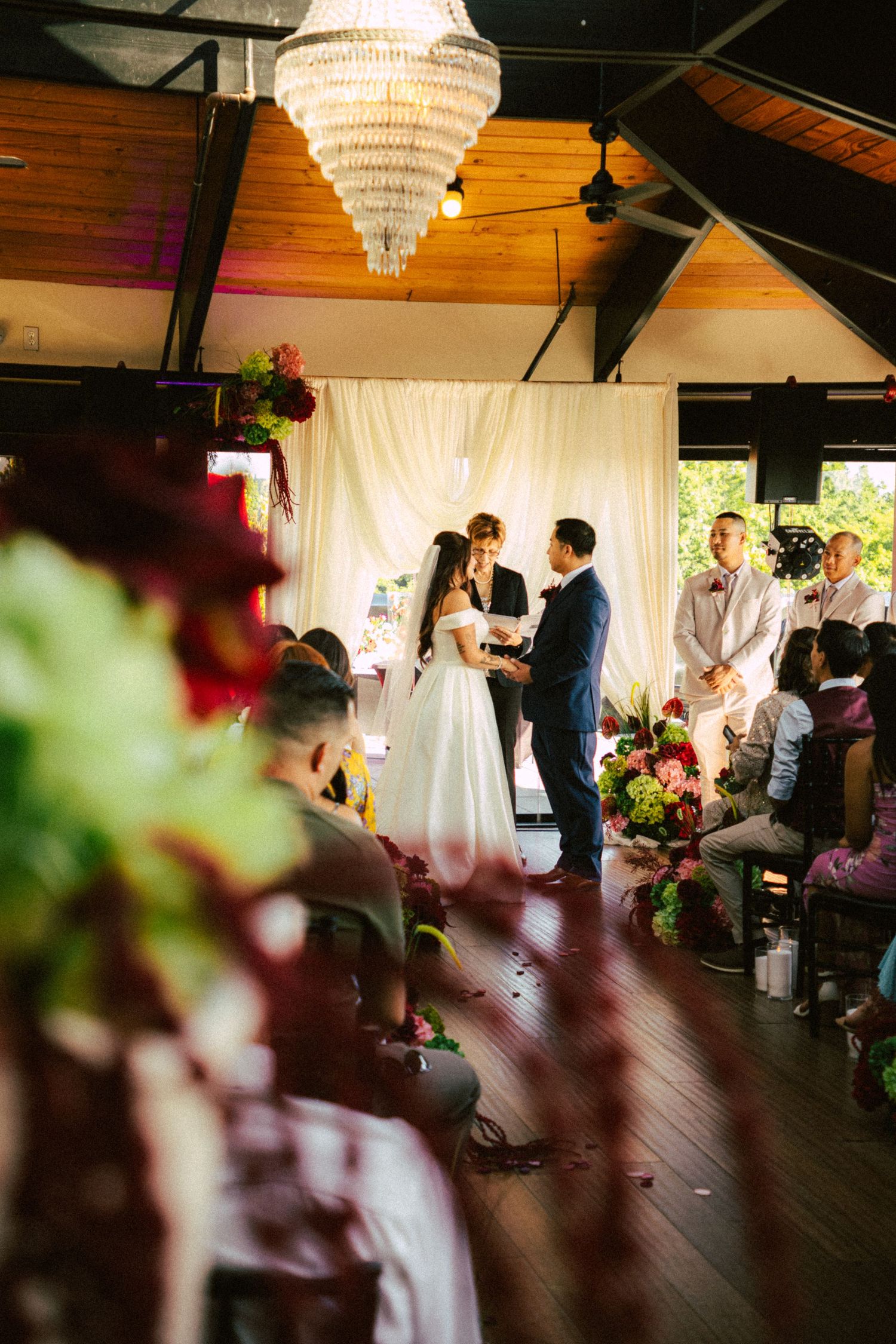 Bride and groom exchange vows at altar beneath chandelier surrounded by lush floral arrangements and guests.