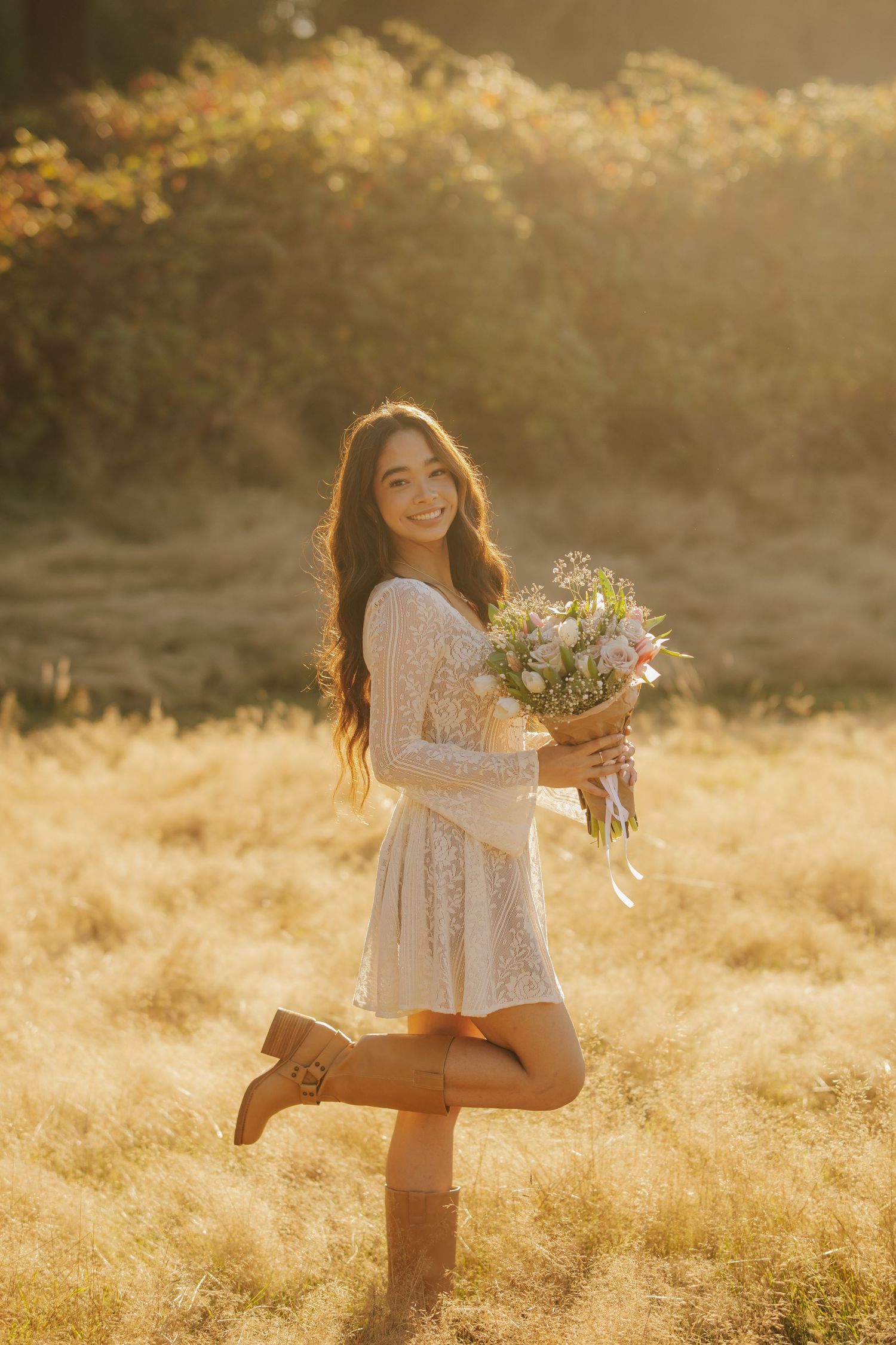 Smiling woman in a white lace dress and brown boots holds a pastel flower bouquet in a golden field at sunset.
