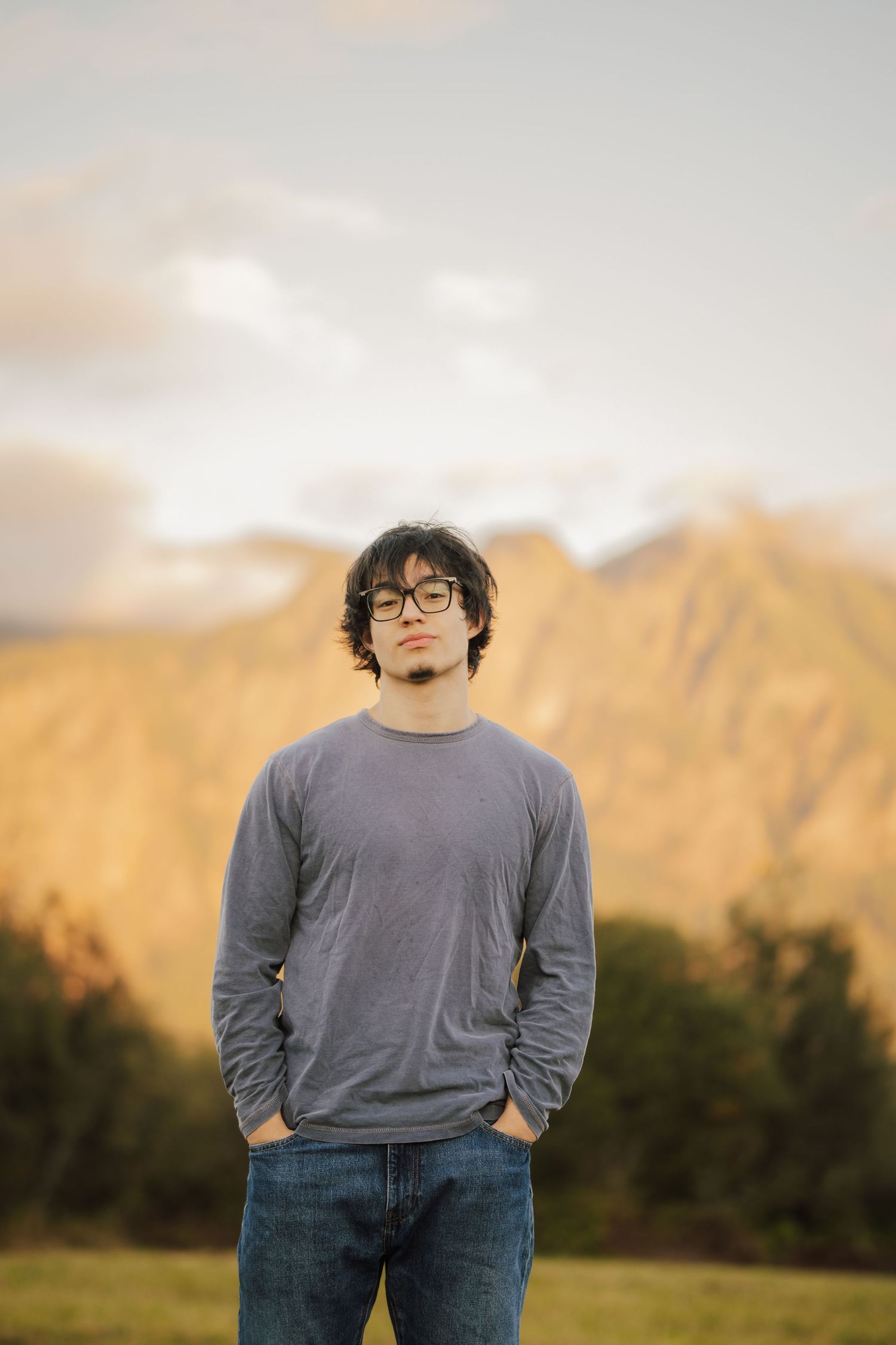 Young man with glasses and curly dark hair stands with hands in pockets in a grassy field before golden mountains at sunset.