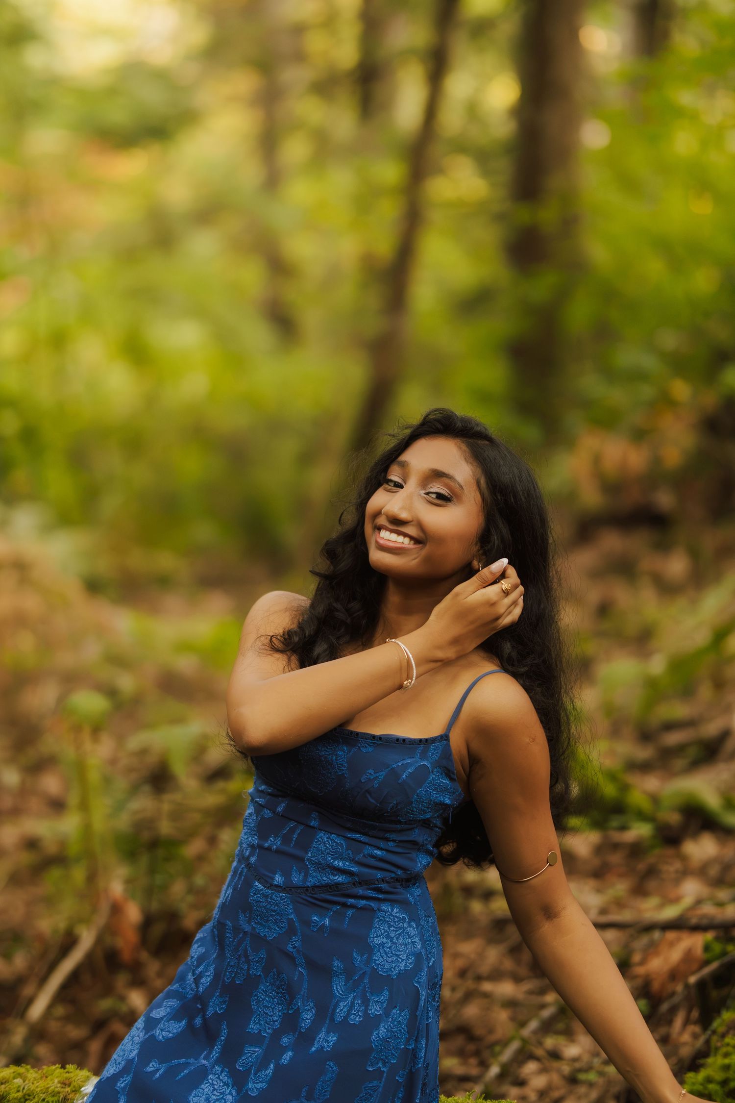 Smiling woman with long dark hair in a blue floral dress poses in a sunlit forest, one hand touching her hair.