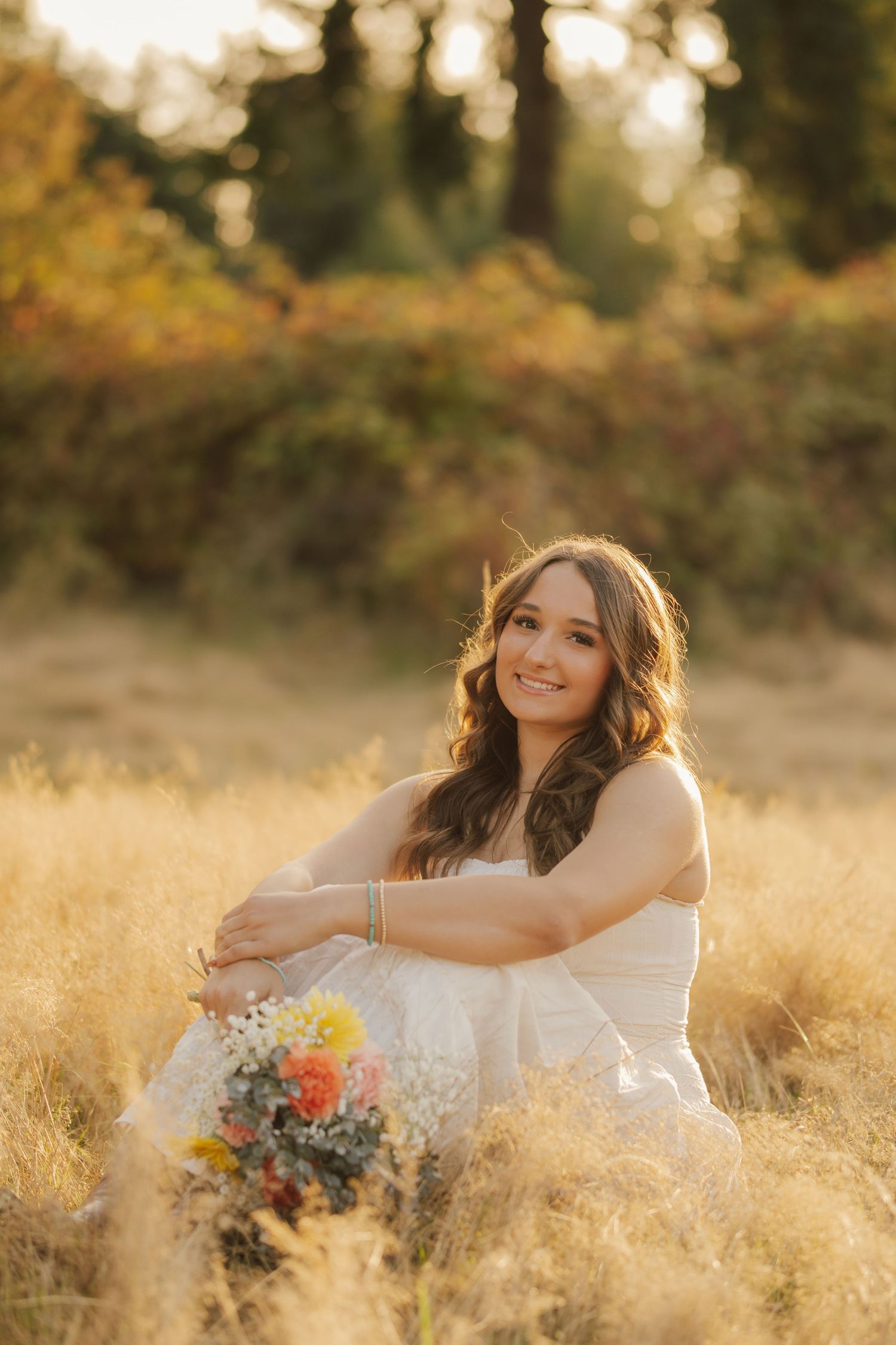 Smiling woman in a white dress sits in golden tall grass holding a colorful flower bouquet, with warm autumn foliage glowing behind her.