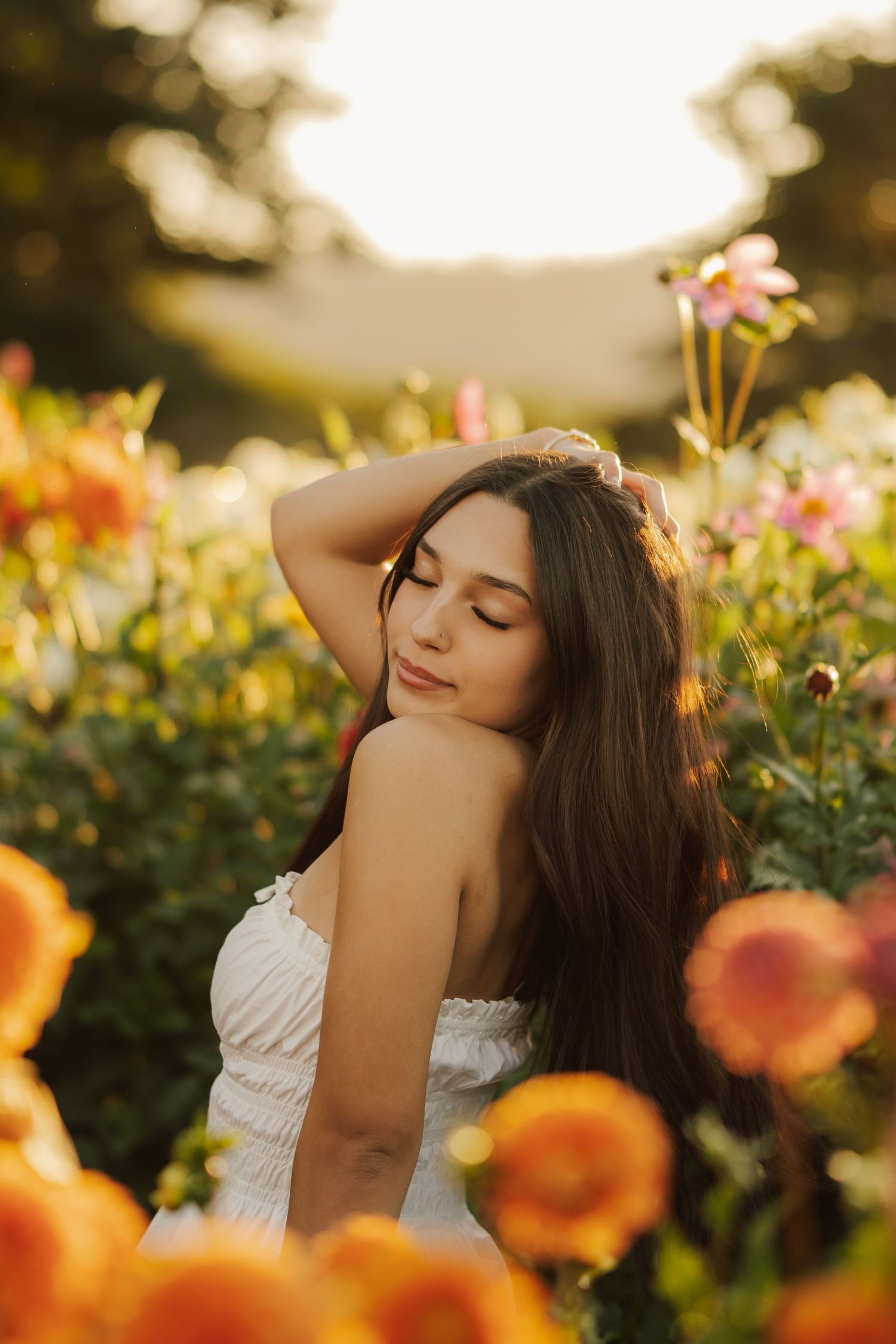 Young woman with long dark hair and closed eyes poses in a golden-hour flower field surrounded by orange and pink blooms.