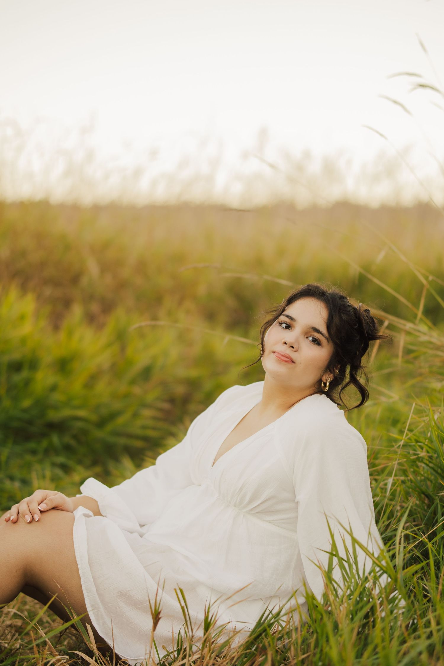 Young woman with dark curly updo in a white V-neck dress sitting in tall golden-green grass during a soft sunset portrait session.