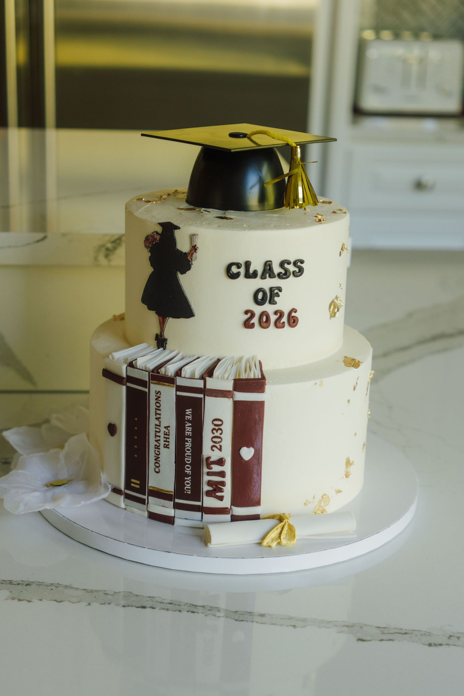 Two-tier white graduation cake with MIT books and Class of 2026 cap topper decoration.
