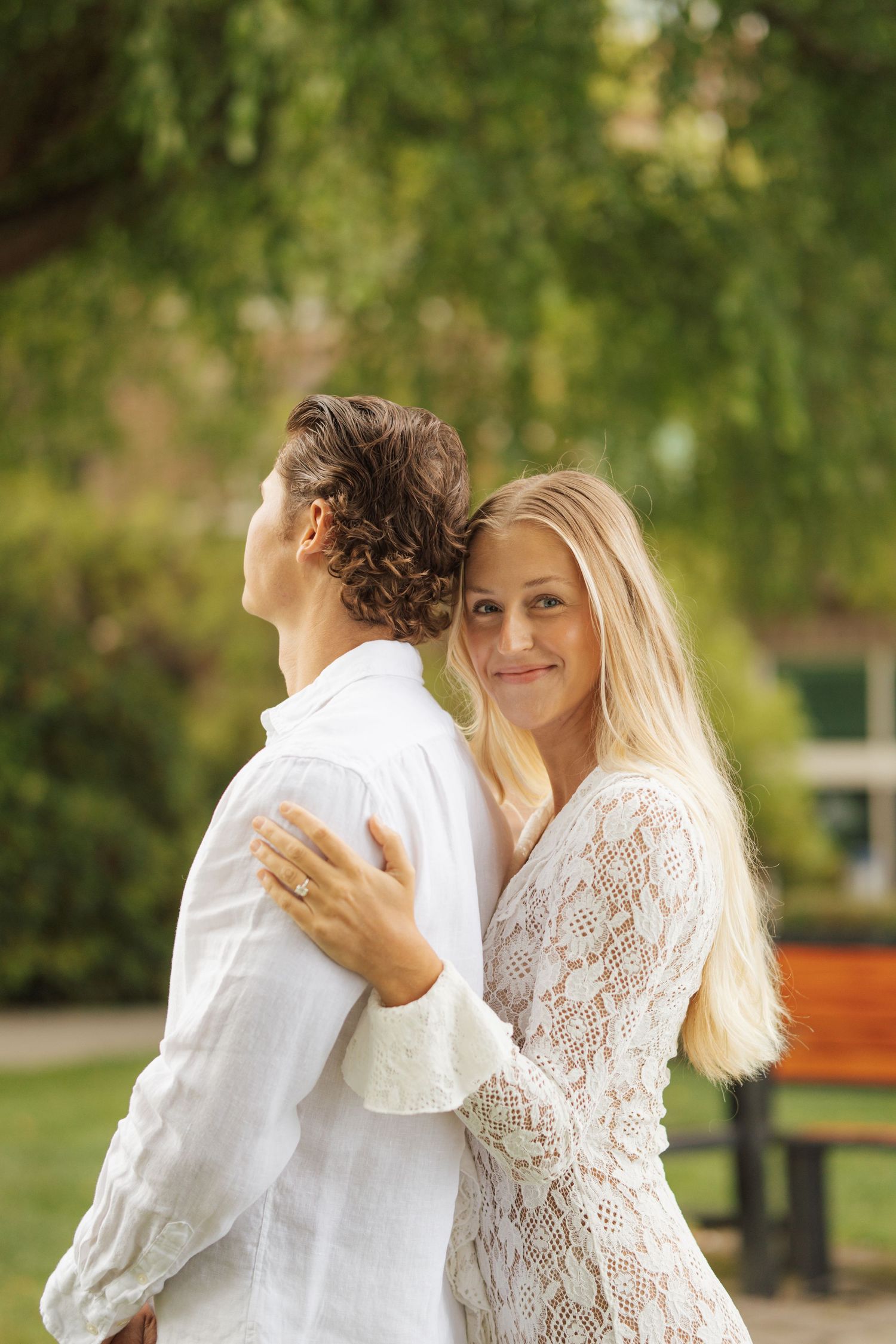 A smiling blonde woman in white lace dress embraces a man in a sunny green park.
