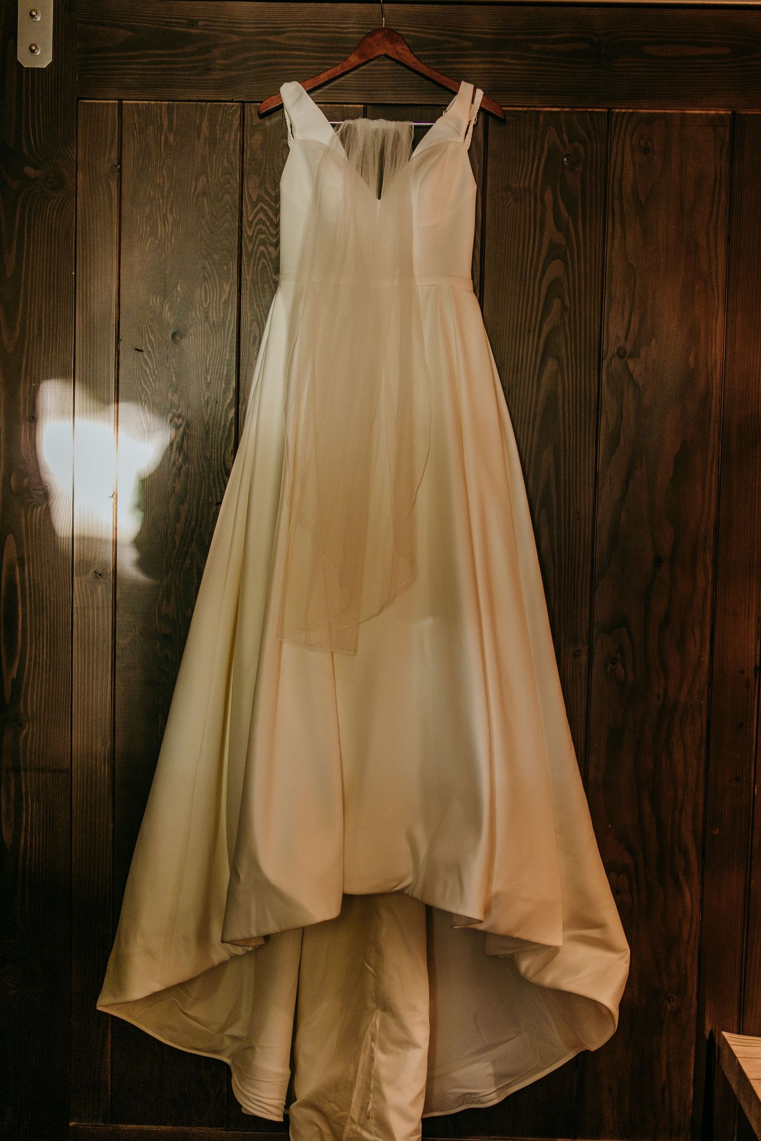 Elegant ivory A-line wedding gown with V-neckline hanging on wooden wall in warm light.