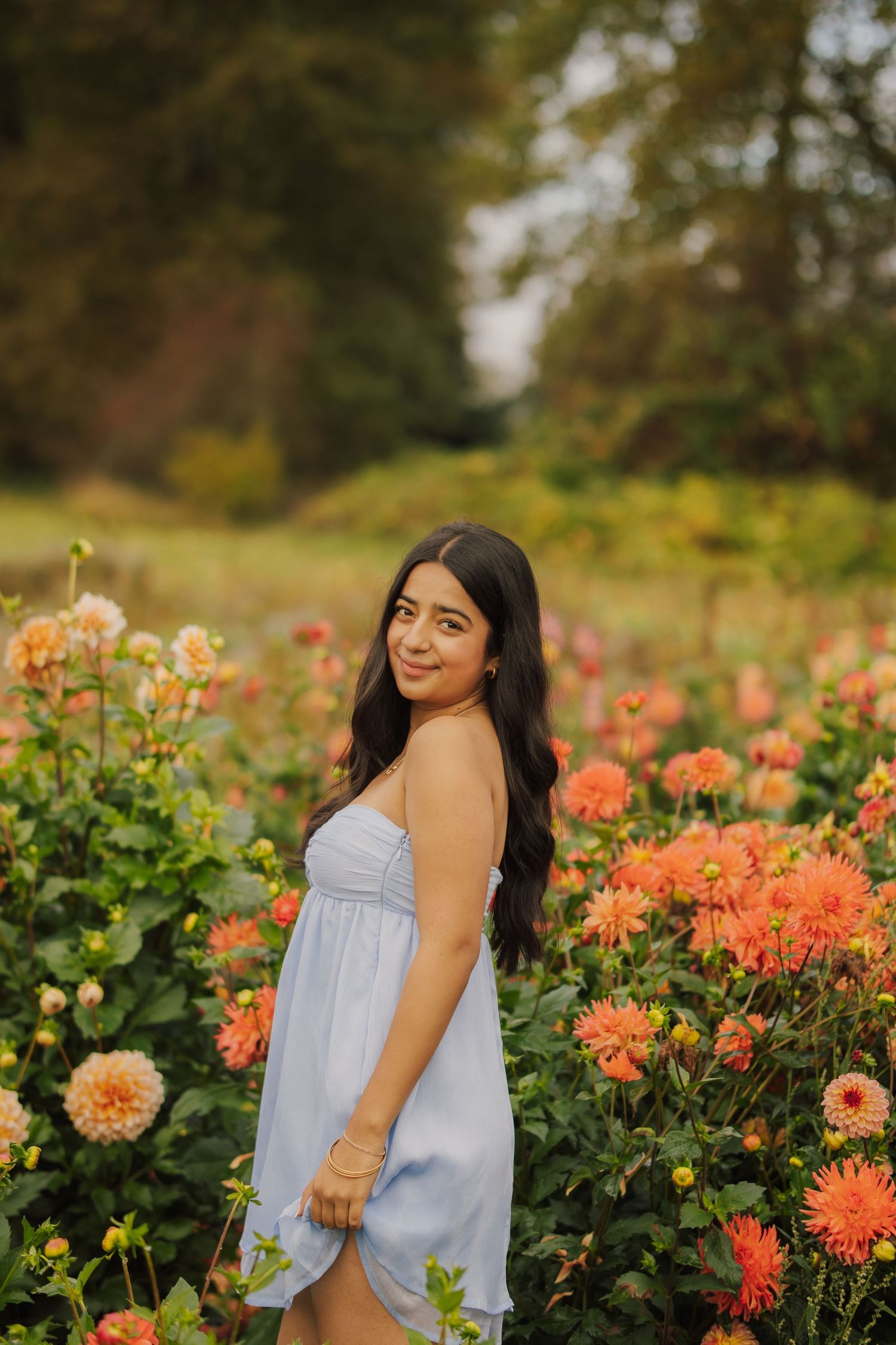 Young woman with long dark hair in a light blue sundress smiles while standing among blooming orange dahlias in a sunlit flower field.