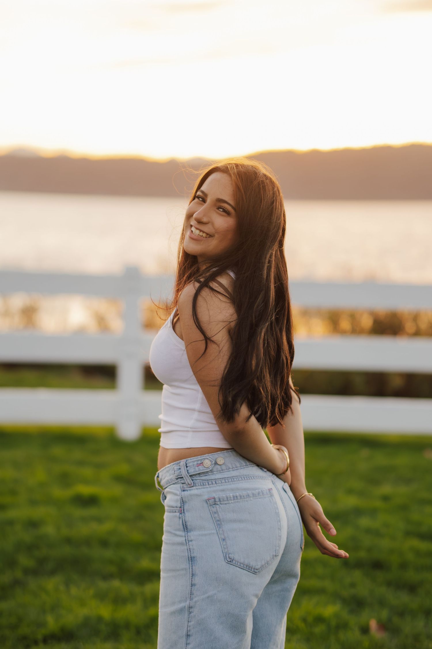 Smiling woman with long brown hair in a white crop top and light-wash jeans glances over her shoulder on a sunset lakeside lawn.