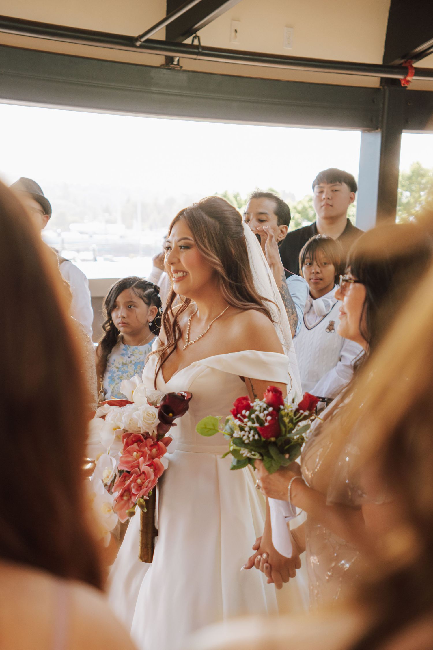 Radiant bride in white veil and off-shoulder gown smiles while holding bouquet surrounded by wedding guests.