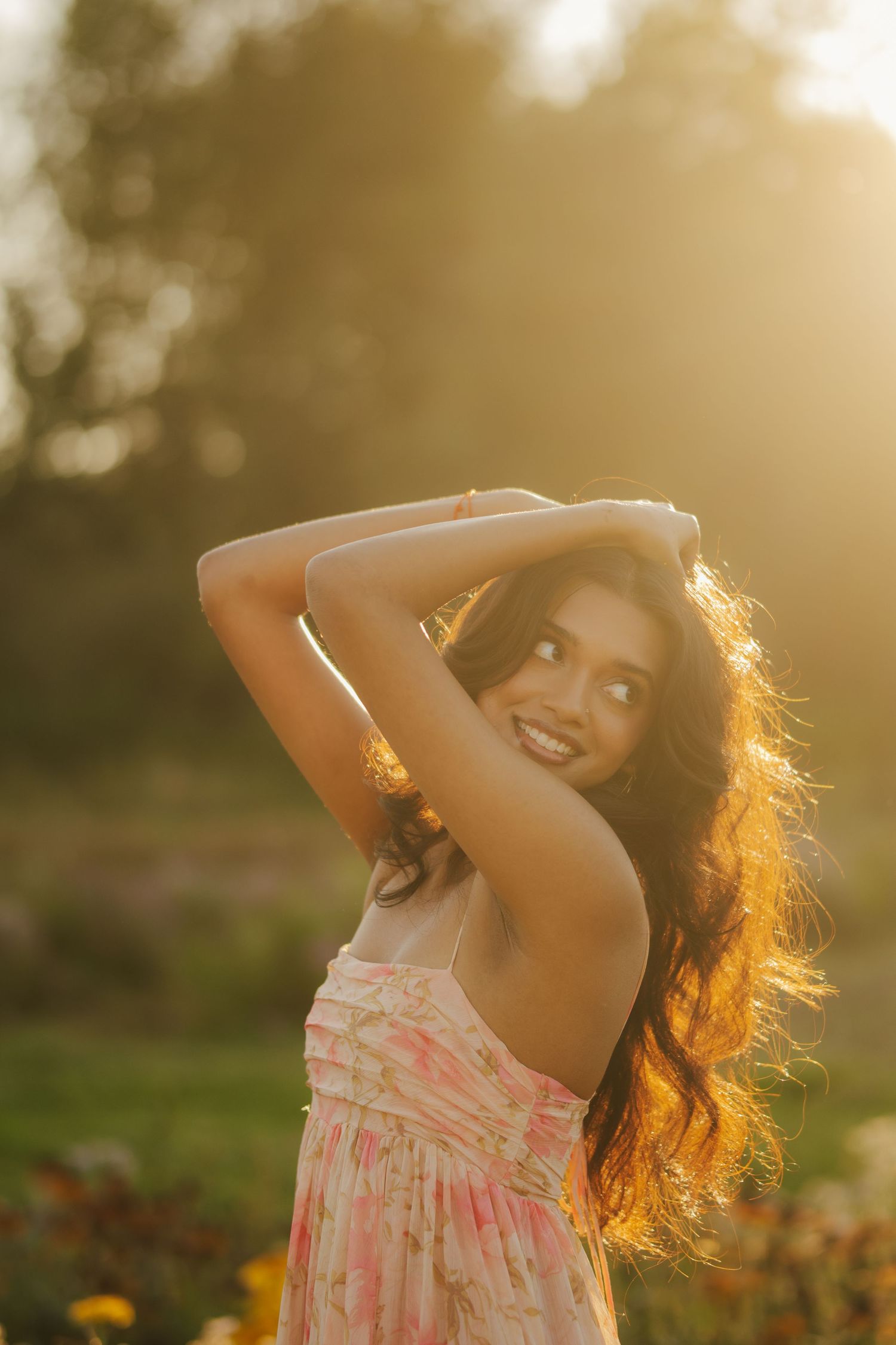 Smiling woman in a pink floral sundress poses with arms raised in a sunlit flower field, her wavy hair glowing in golden hour light.