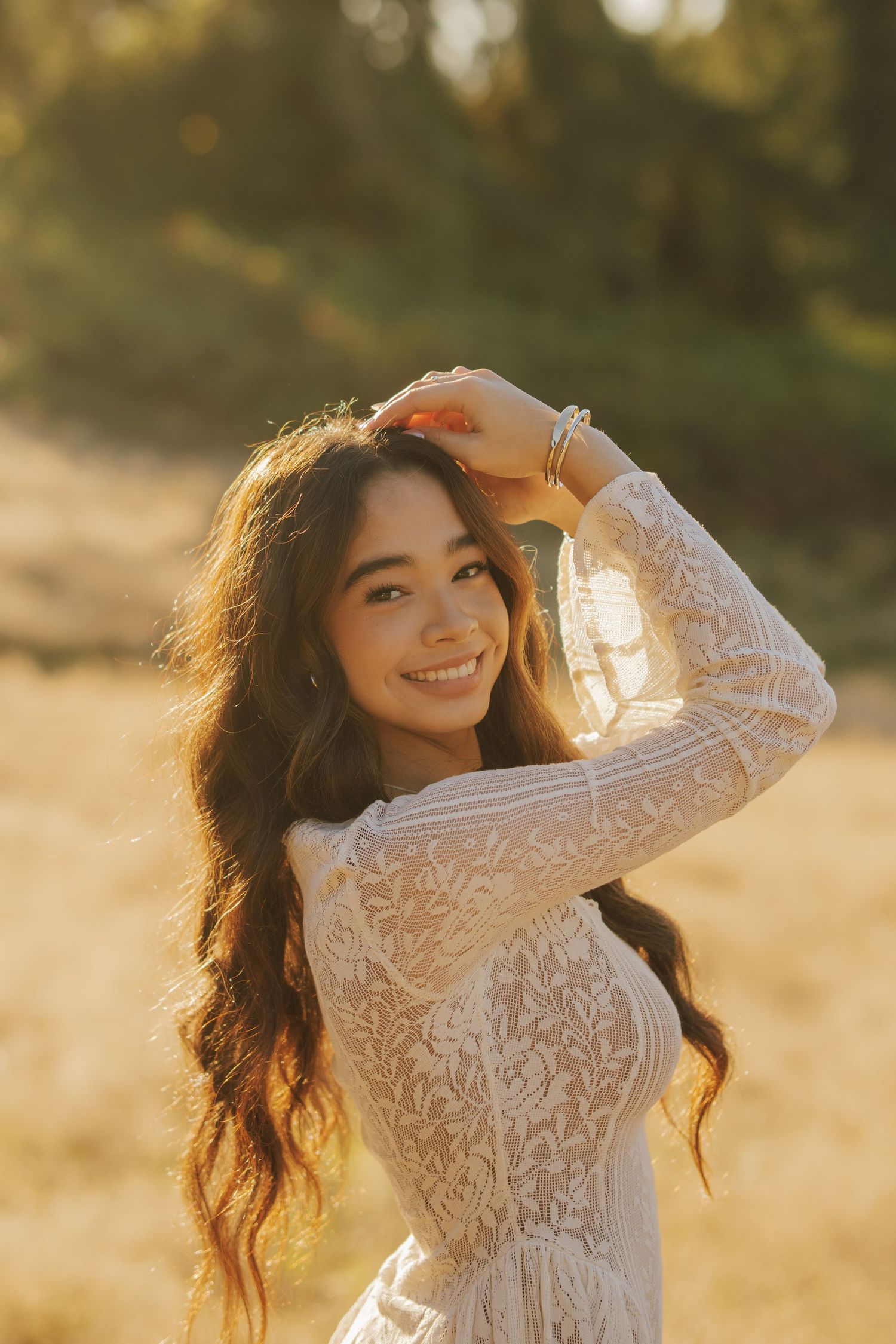 Smiling woman with long wavy brown hair in a white lace dress poses with her hand on her head in warm golden sunlight outdoors.