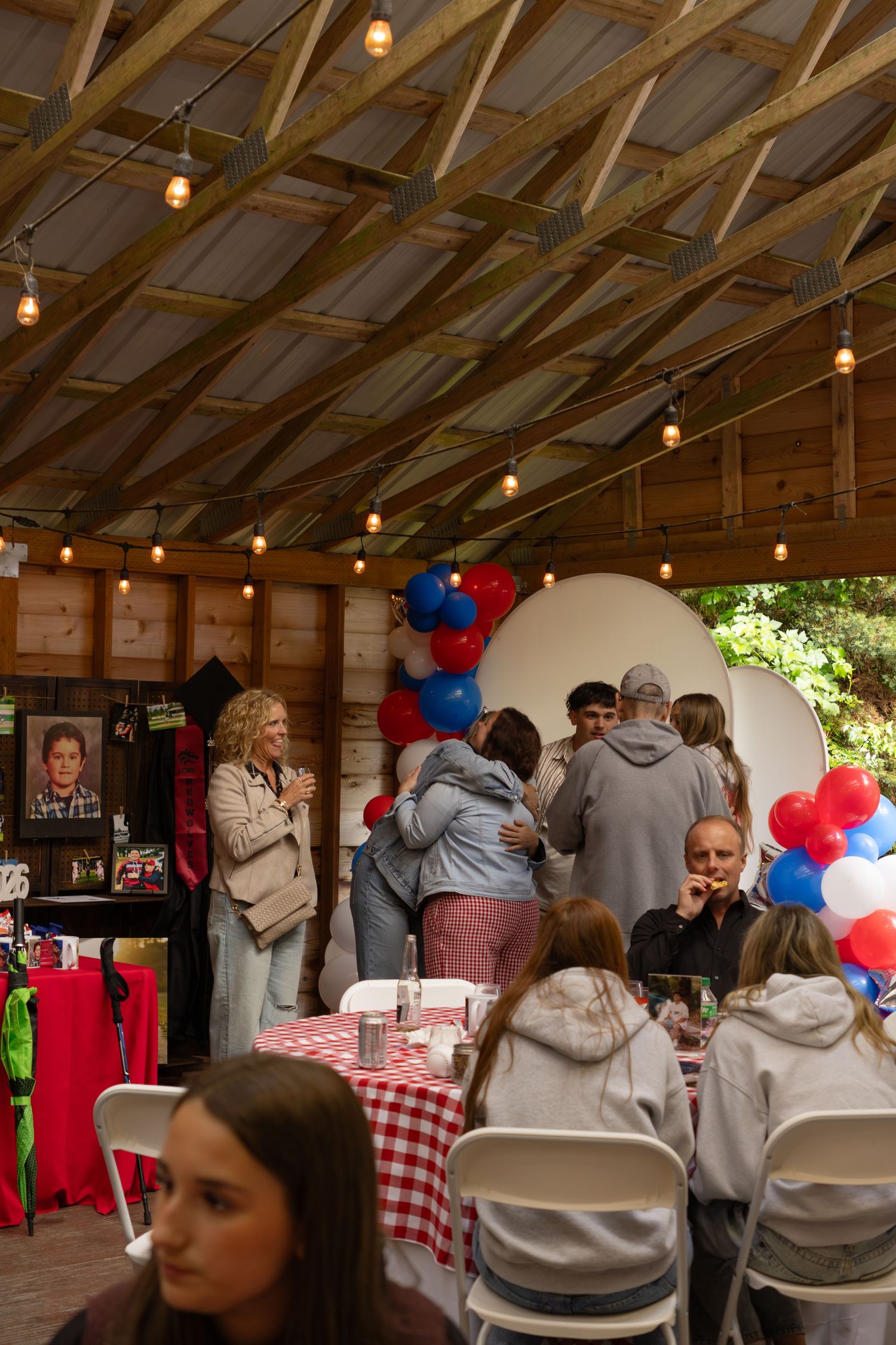 Guests hugging and socializing at a party with red, white, and blue balloon decorations.