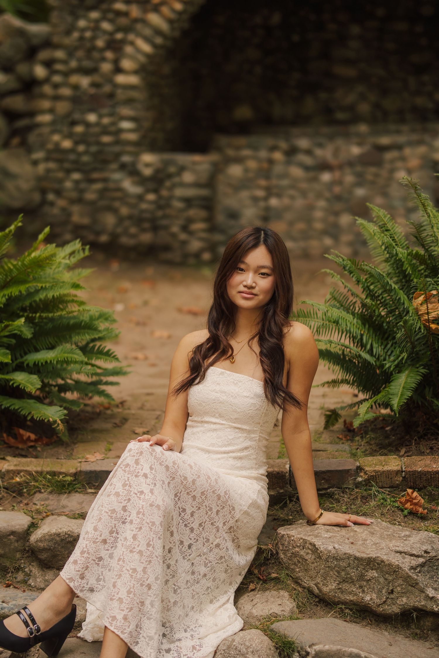 Woman with long dark hair sits on a stone ledge in a white lace dress, surrounded by green ferns and a rustic stone wall.