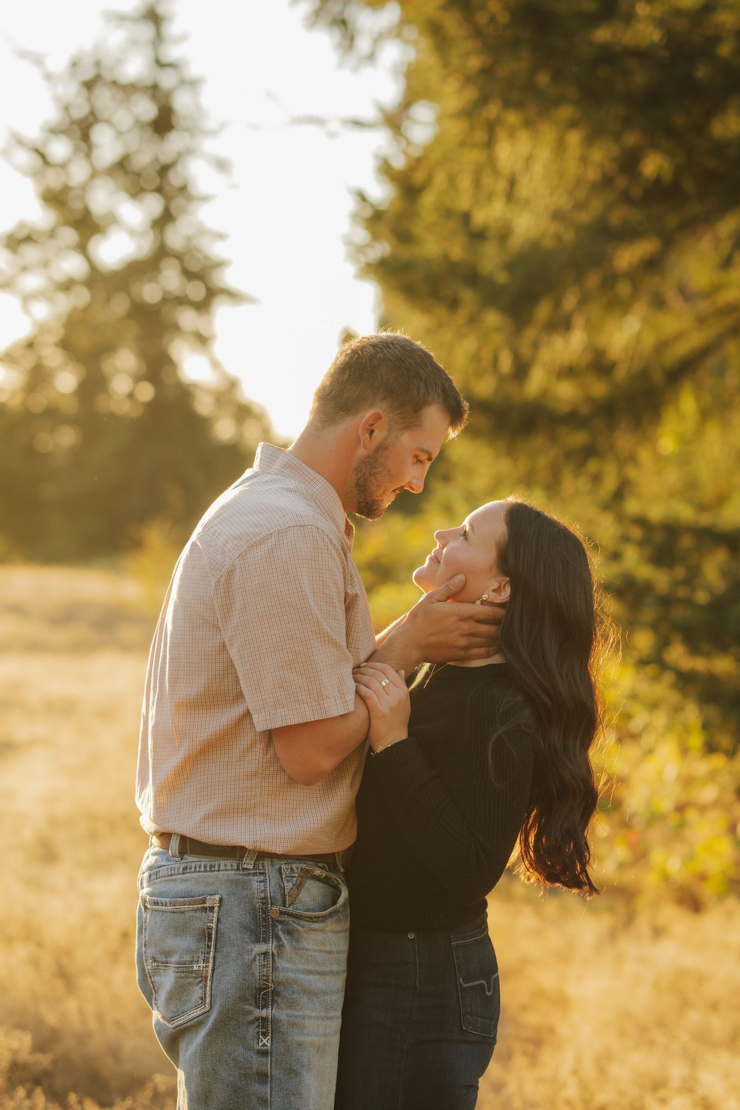 Couple embracing in a golden sunlit field at sunset, man in a tan button-up shirt gently cradling the woman's face before a kiss.