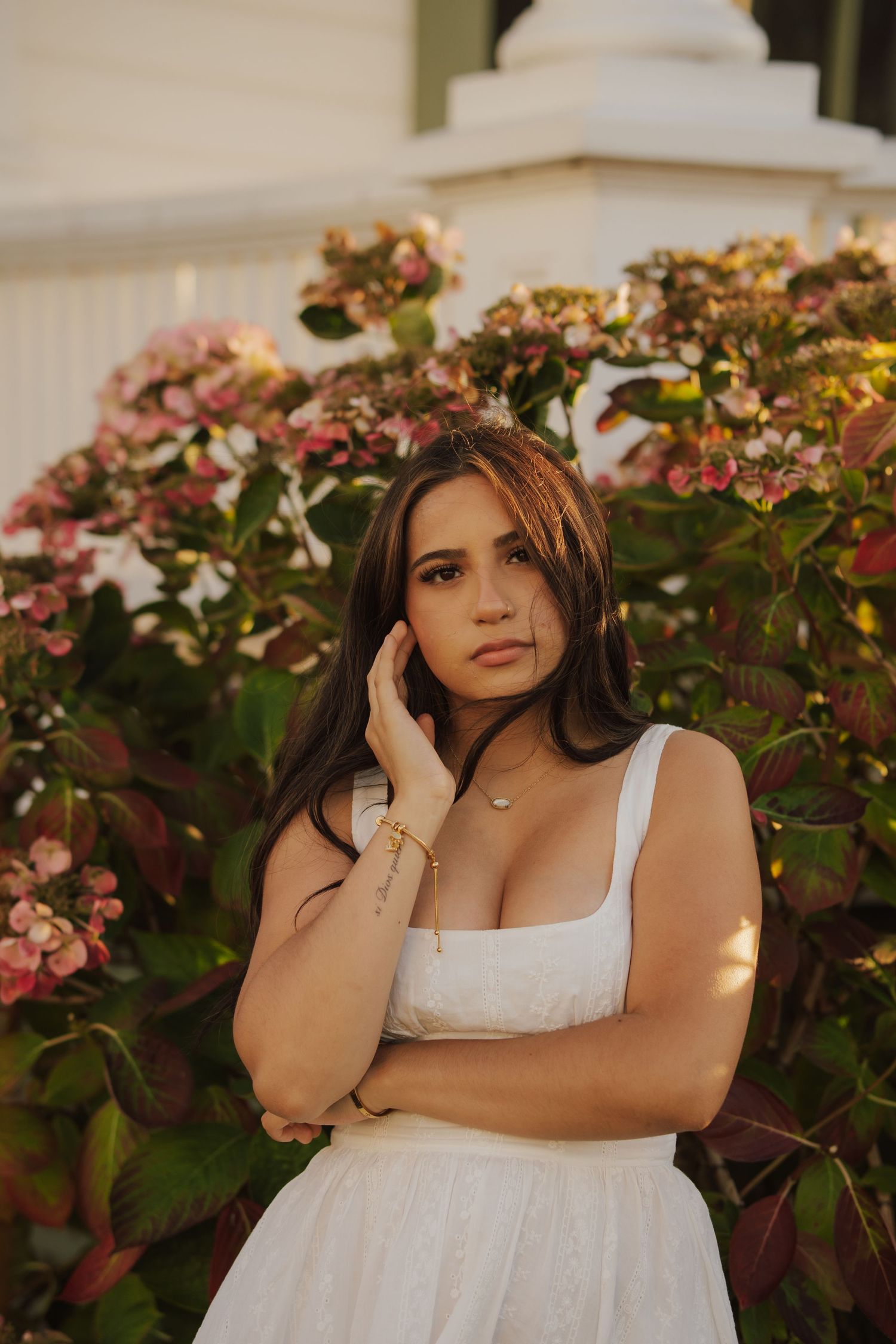 Young woman in a white sundress poses with her hand at her cheek in front of blooming pink hydrangeas in warm golden-hour light.