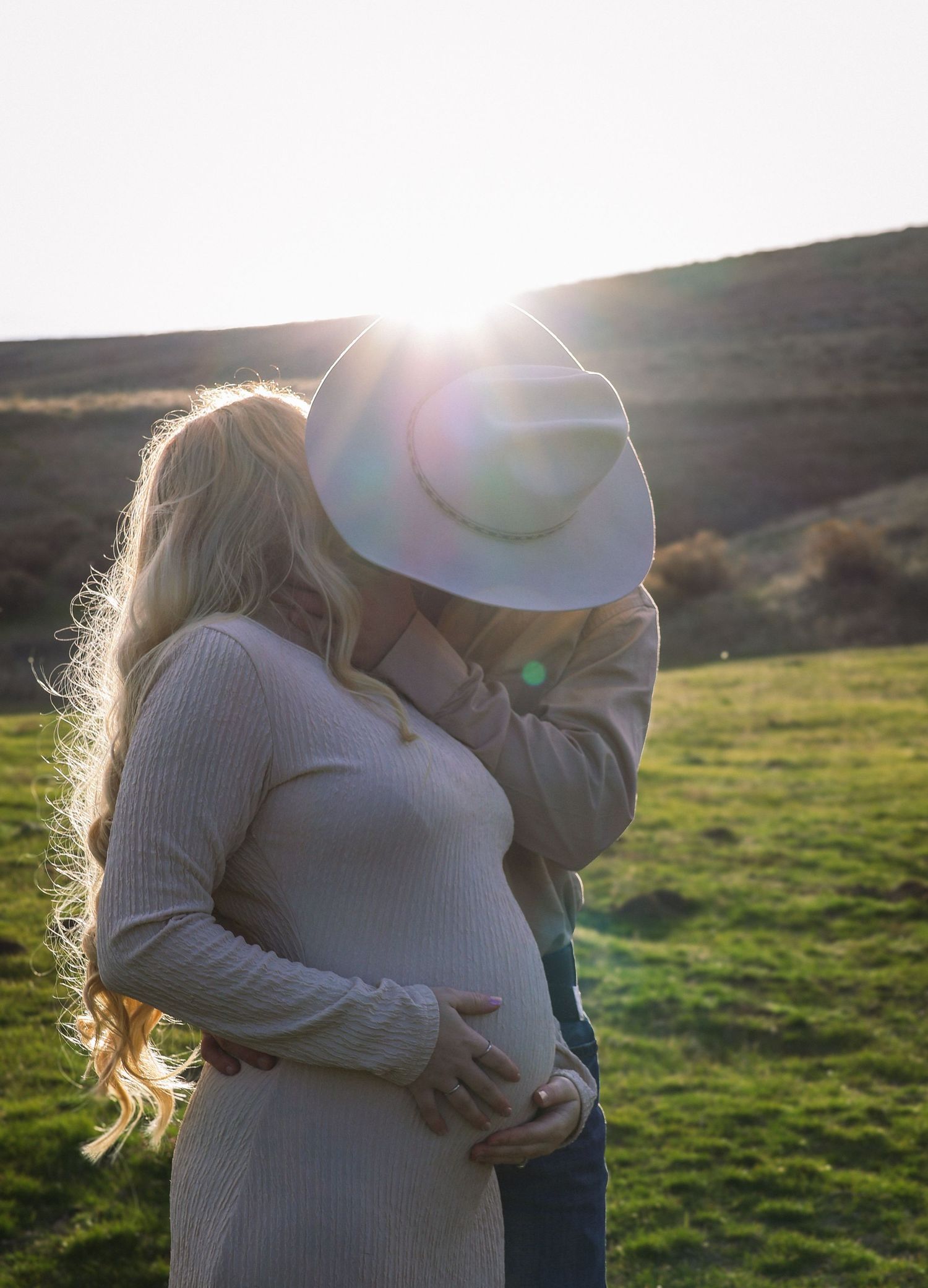 A couple kisses while their faces are hidden behind a cowboy hat, and tenderly holding the baby bump in this western maternity photoshoot.