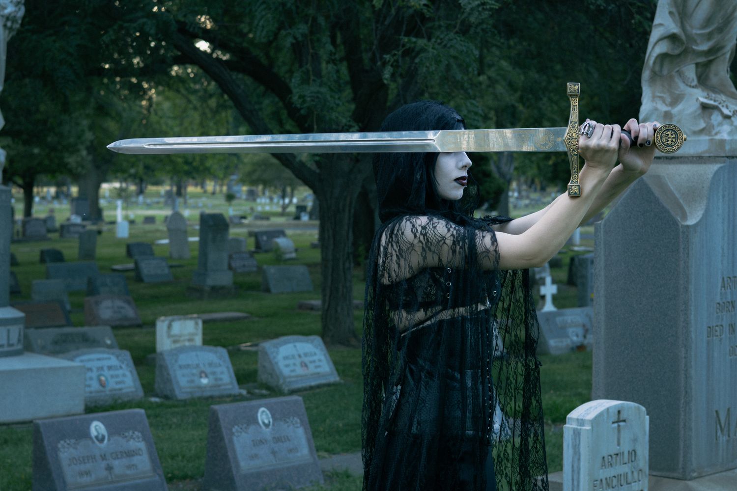 A woman holds a silver sword over her eyes in the graveyard for this halloween photoshoot.