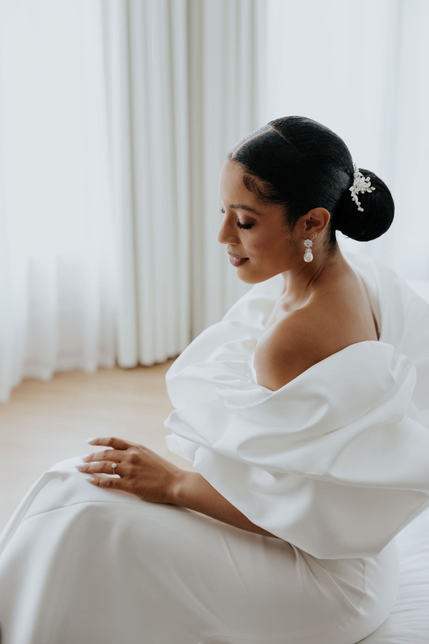 Elegant bride in off-shoulder white satin gown seated by window with updo hairstyle.
