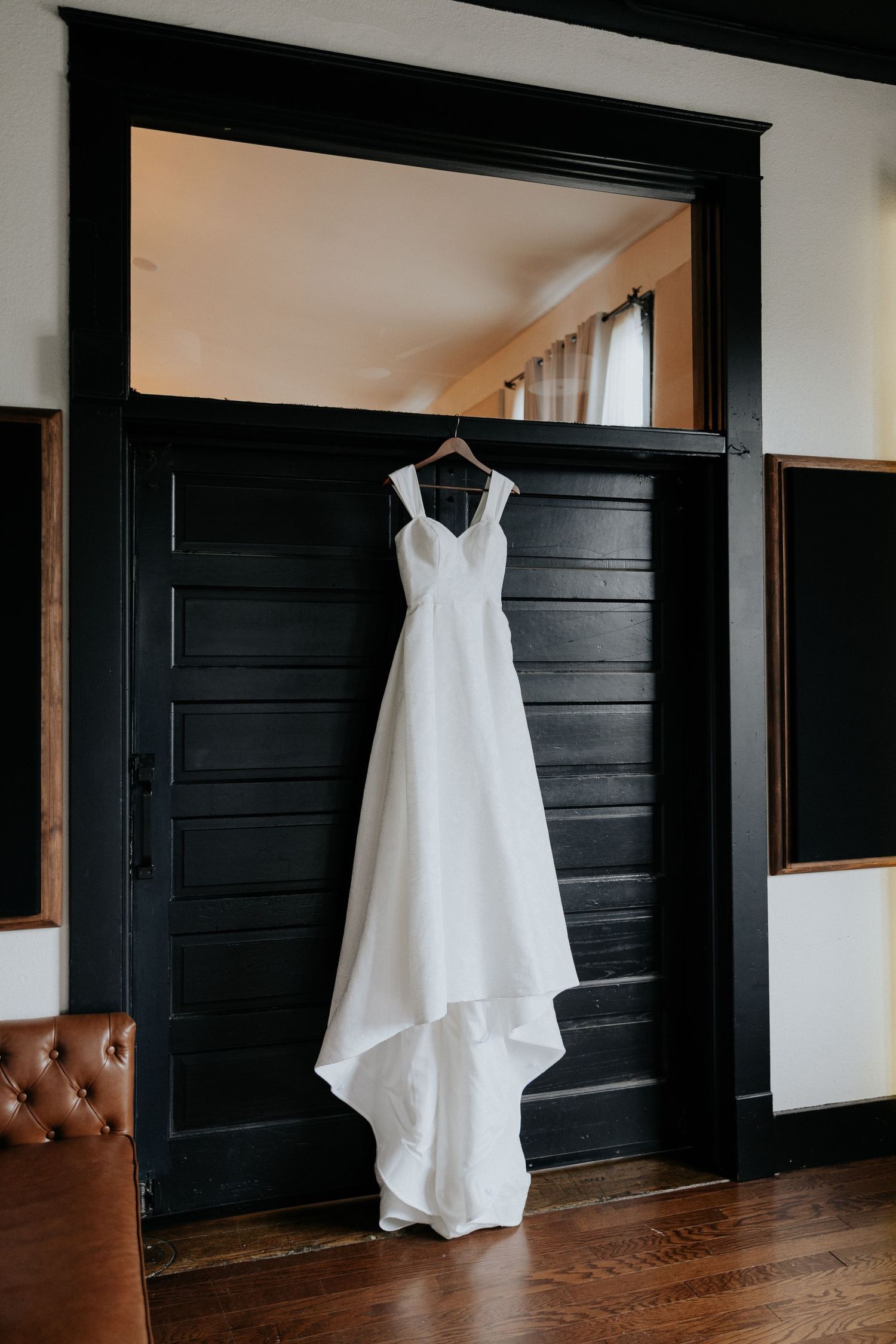 Elegant white A-line wedding dress hanging on black door frame in rustic interior setting.