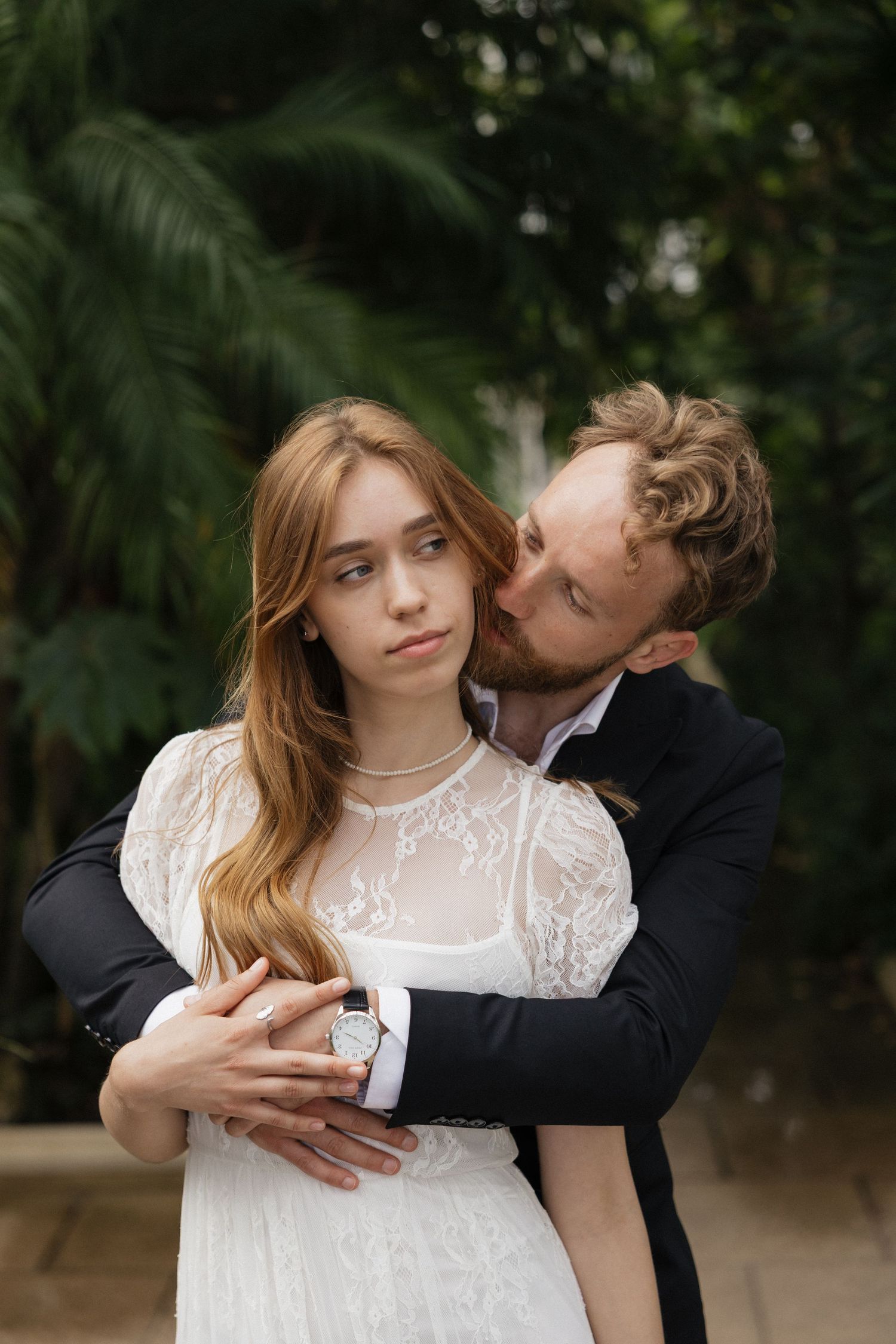 Groom kisses bride on the cheek while embracing her among lush tropical greenery.