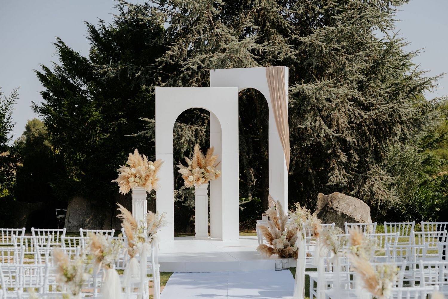 Elegant outdoor wedding ceremony setup with white arch, pampas grass arrangements, chiavari chairs, and white aisle runner.