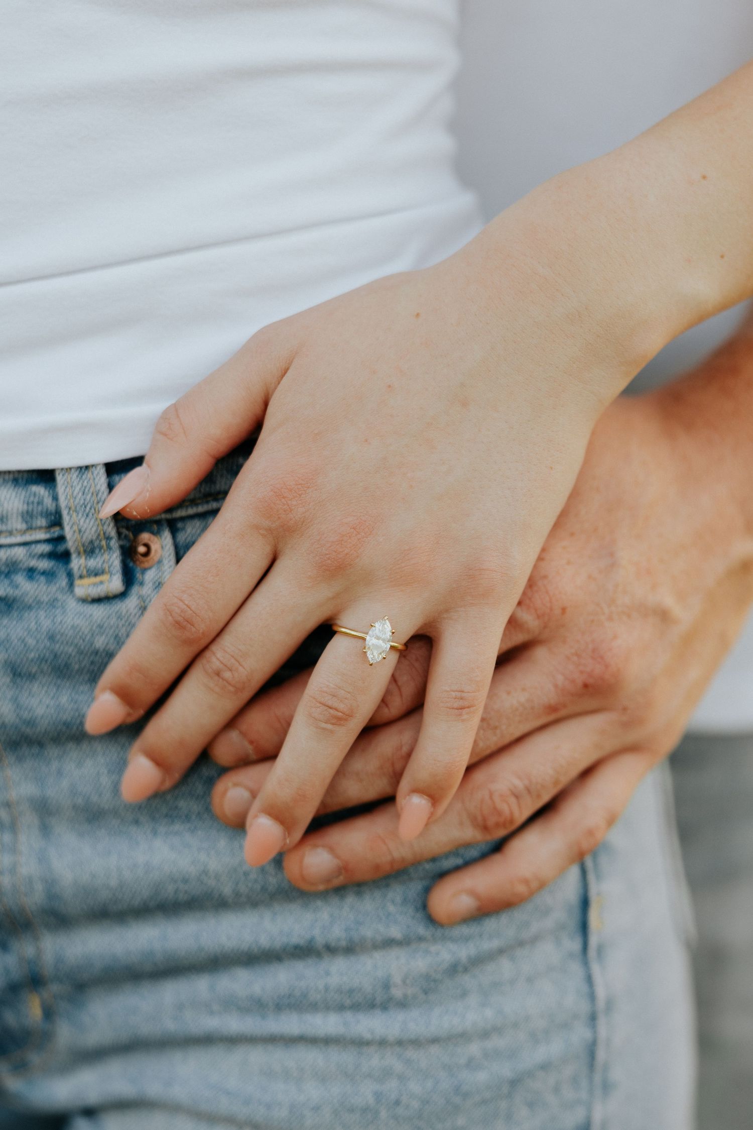 Close-up of woman's hand showing marquise diamond engagement ring resting on partner's hands.