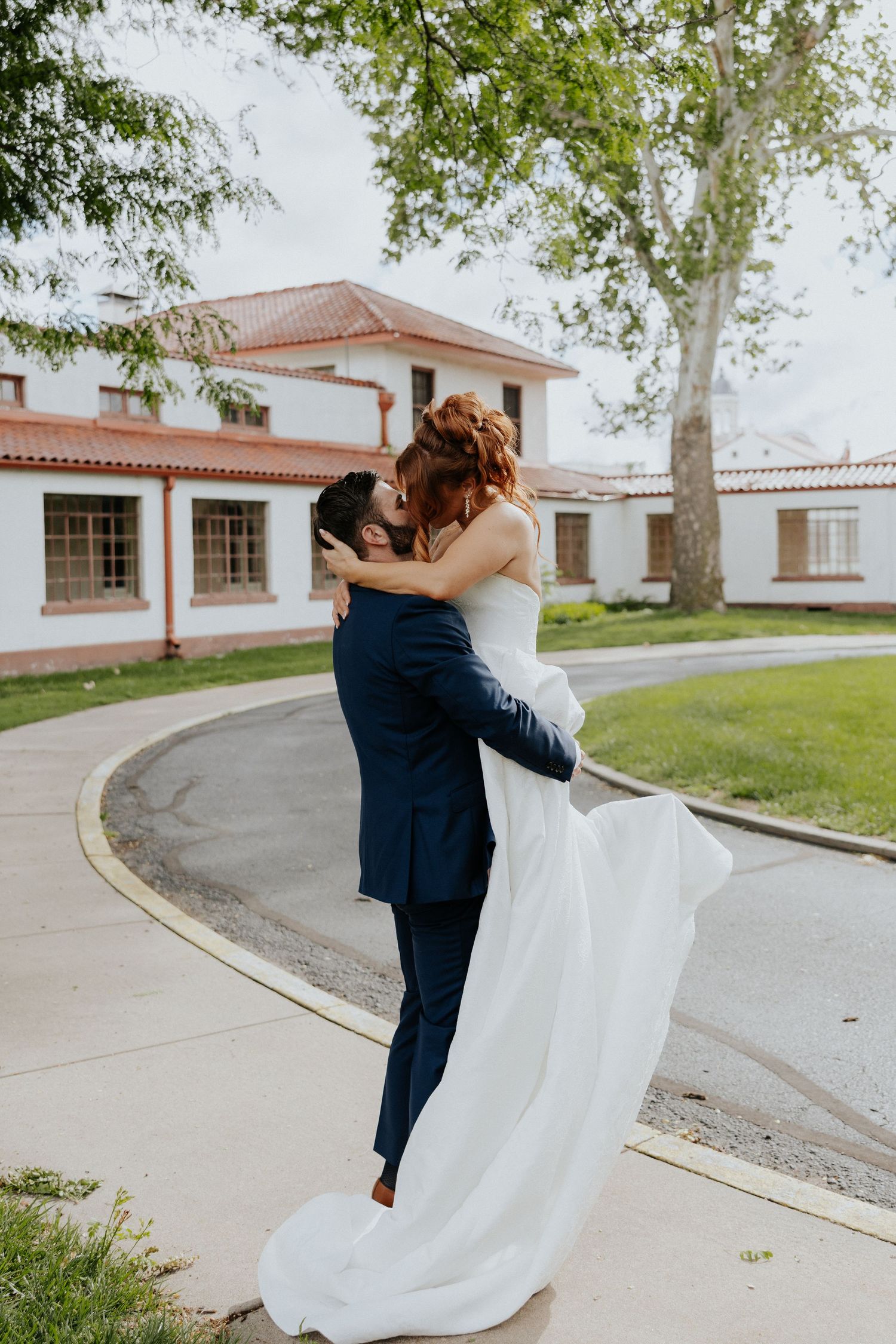 Groom in navy suit lifts bride in white gown as they kiss outdoors near a white building with red tile roof.