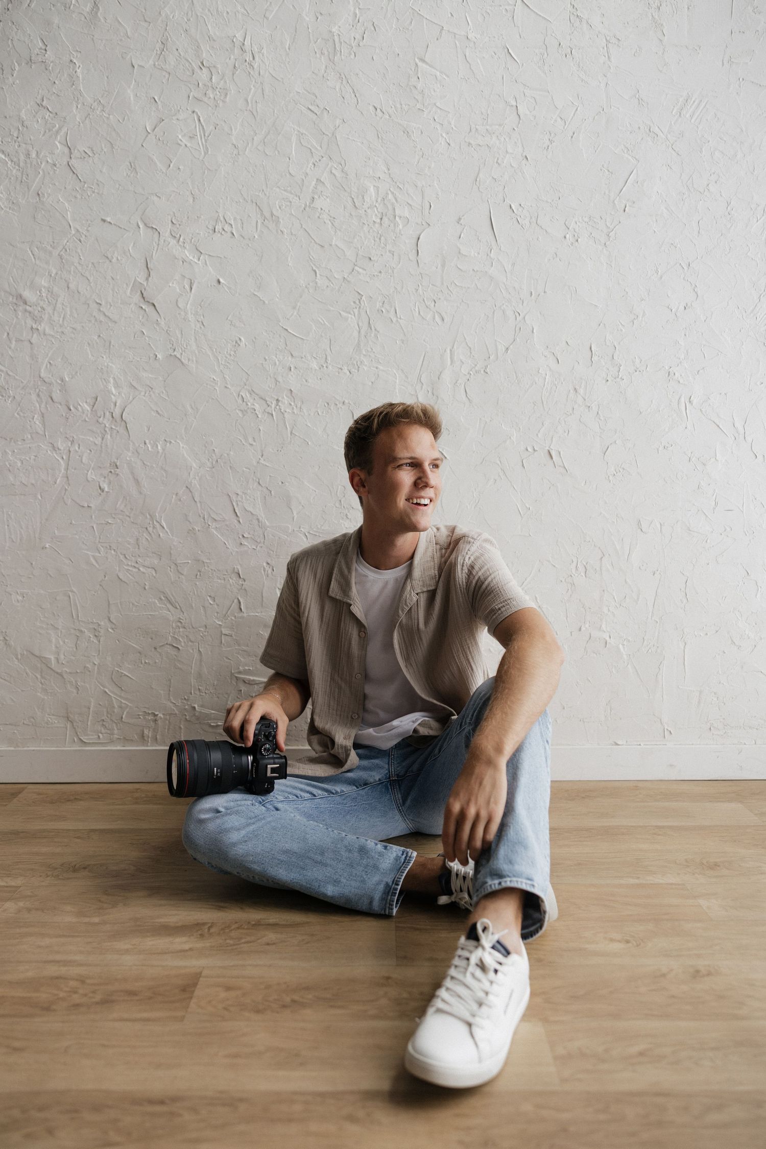 Young male photographer sitting cross-legged on wooden floor holding DSLR camera against white textured wall.