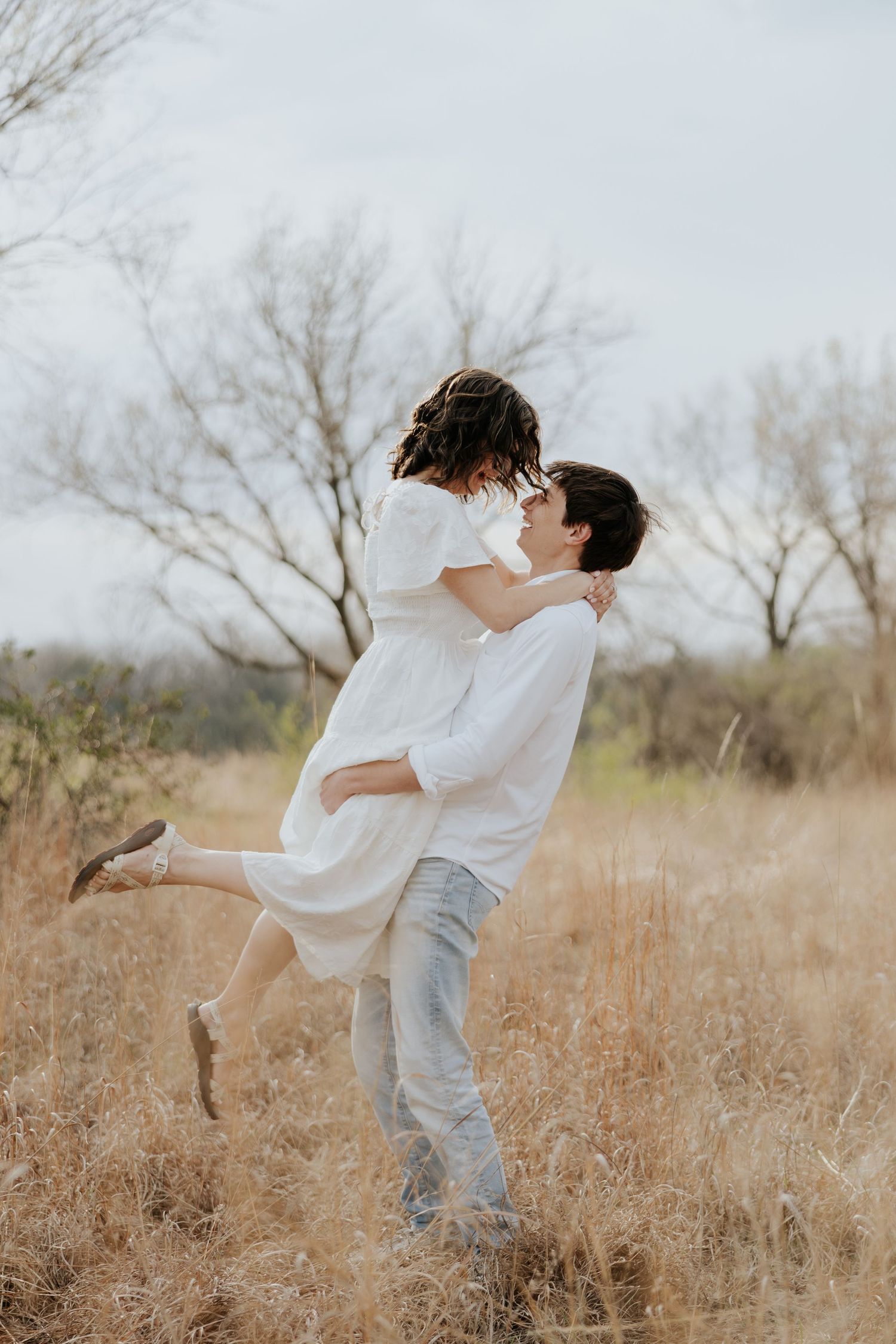 Man lifting woman in white dress in a romantic embrace in dry grass field.