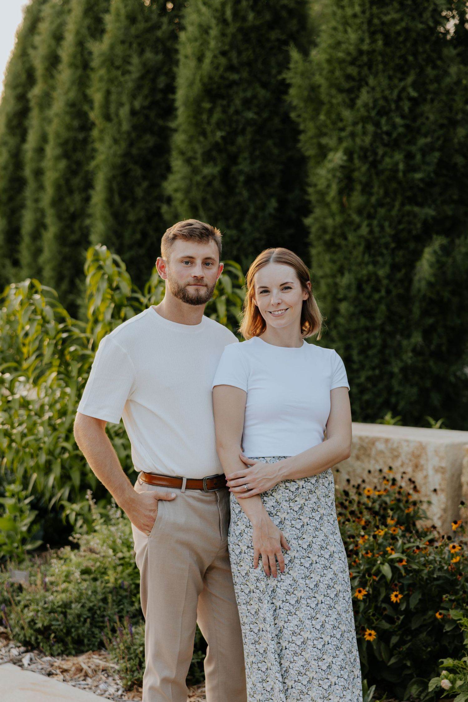 Smiling couple in matching white tops poses together in a sunlit garden with tall evergreens.