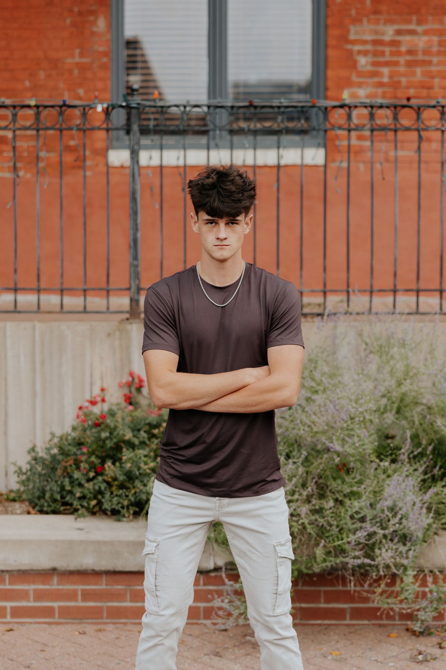 Confident young man in brown t-shirt and gray cargo pants stands with arms crossed before brick wall.