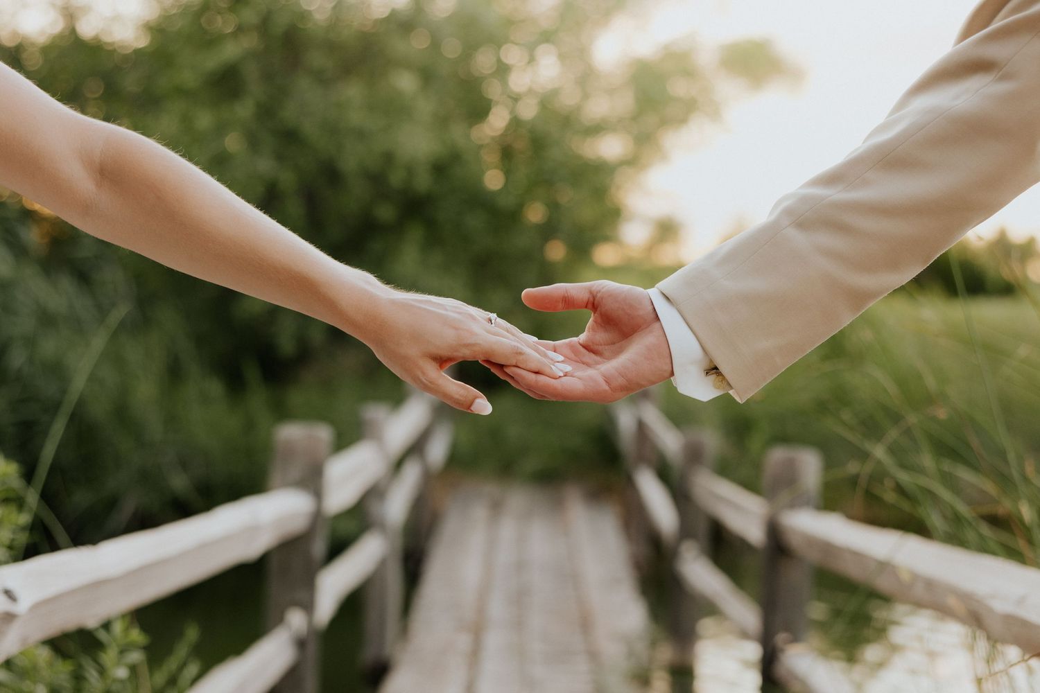 Bride and groom reaching for each other's hands on a wooden bridge surrounded by lush greenery.