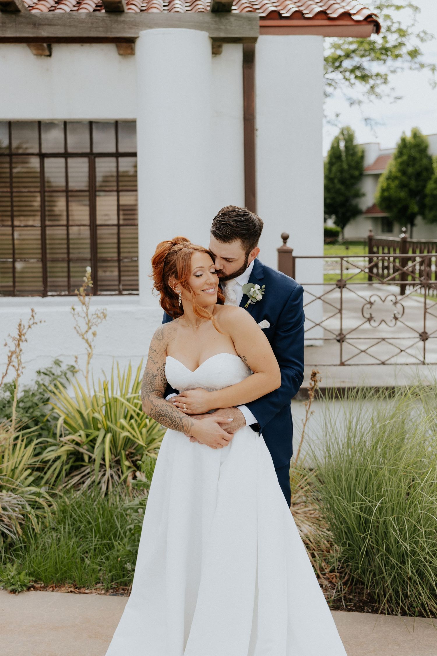 Groom in navy suit embracing tattooed red-haired bride in strapless white gown outdoors.