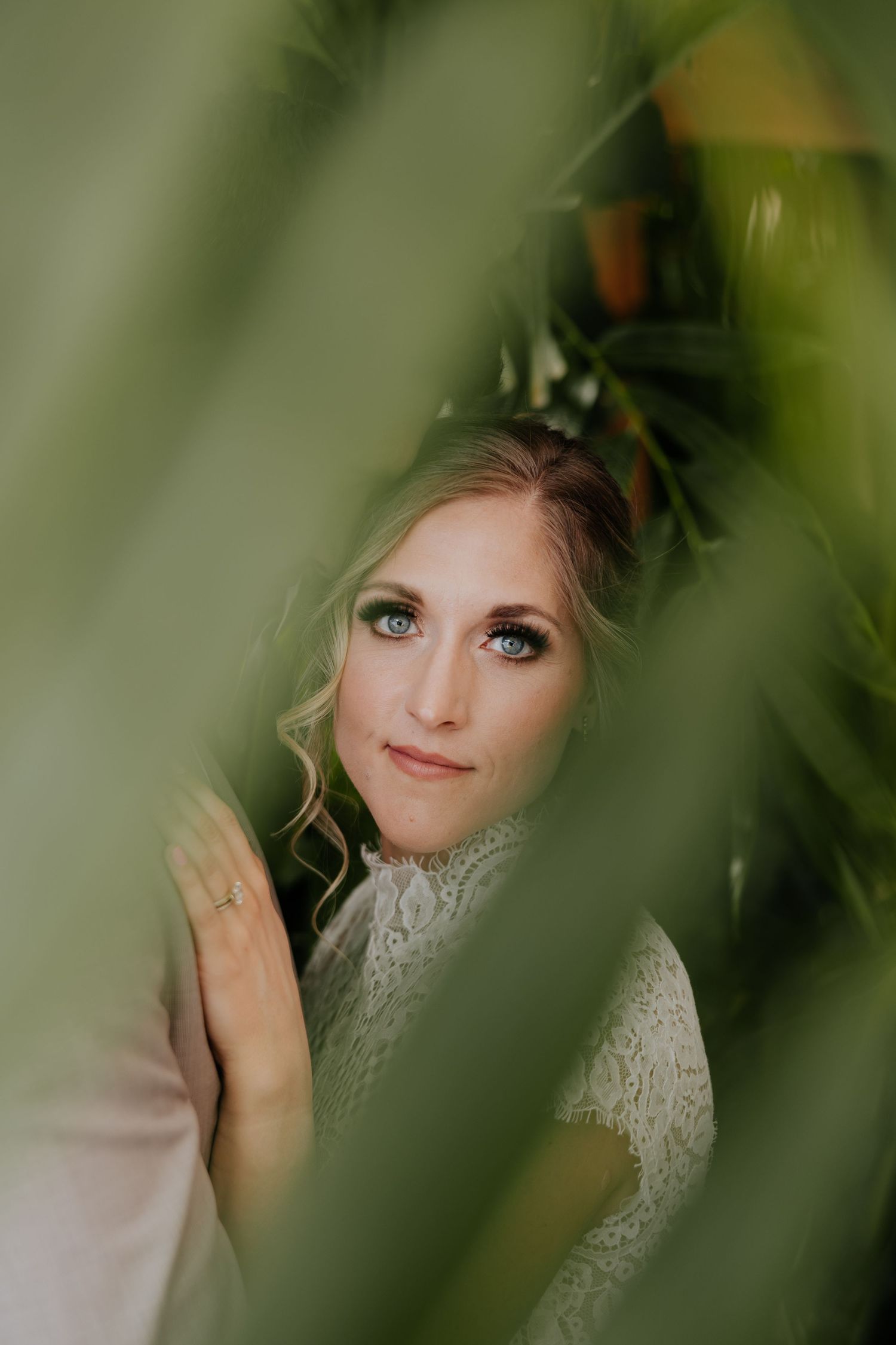 Beautiful blue-eyed bride in lace dress peers through lush tropical green leaves for wedding portrait.