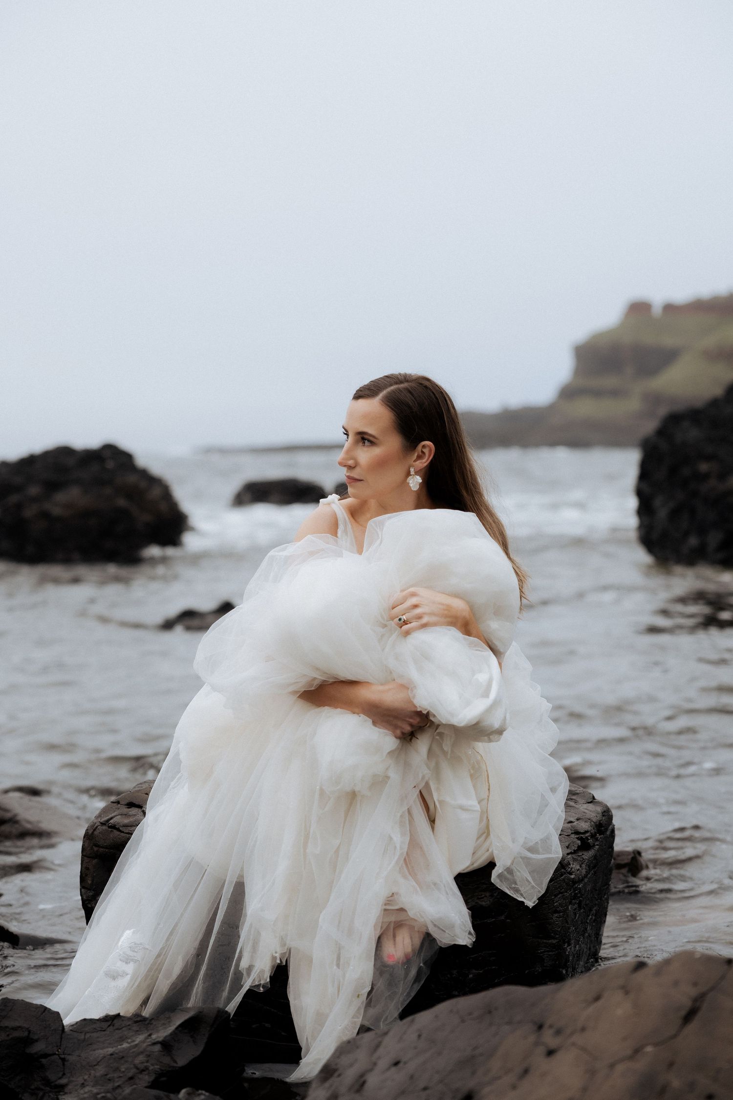 Bride in white tulle gown sitting on dark coastal rocks, gazing serenely at the misty ocean.