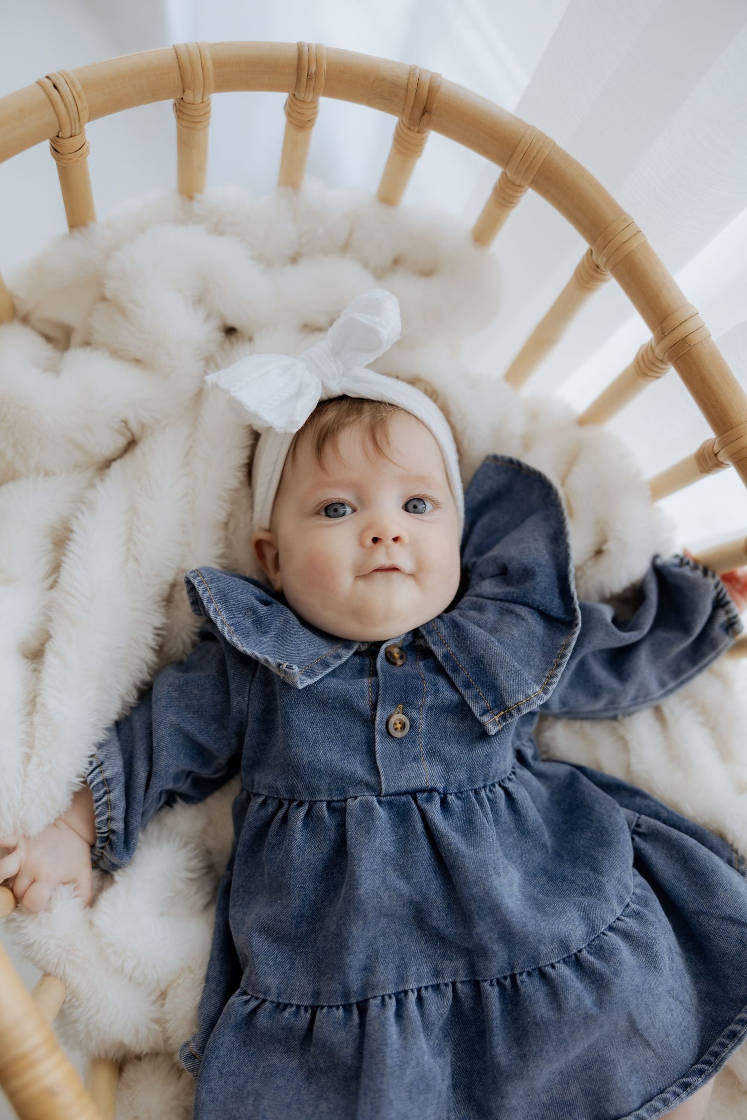 Adorable baby girl in denim dress with white bow headband lies in a wicker bassinet.