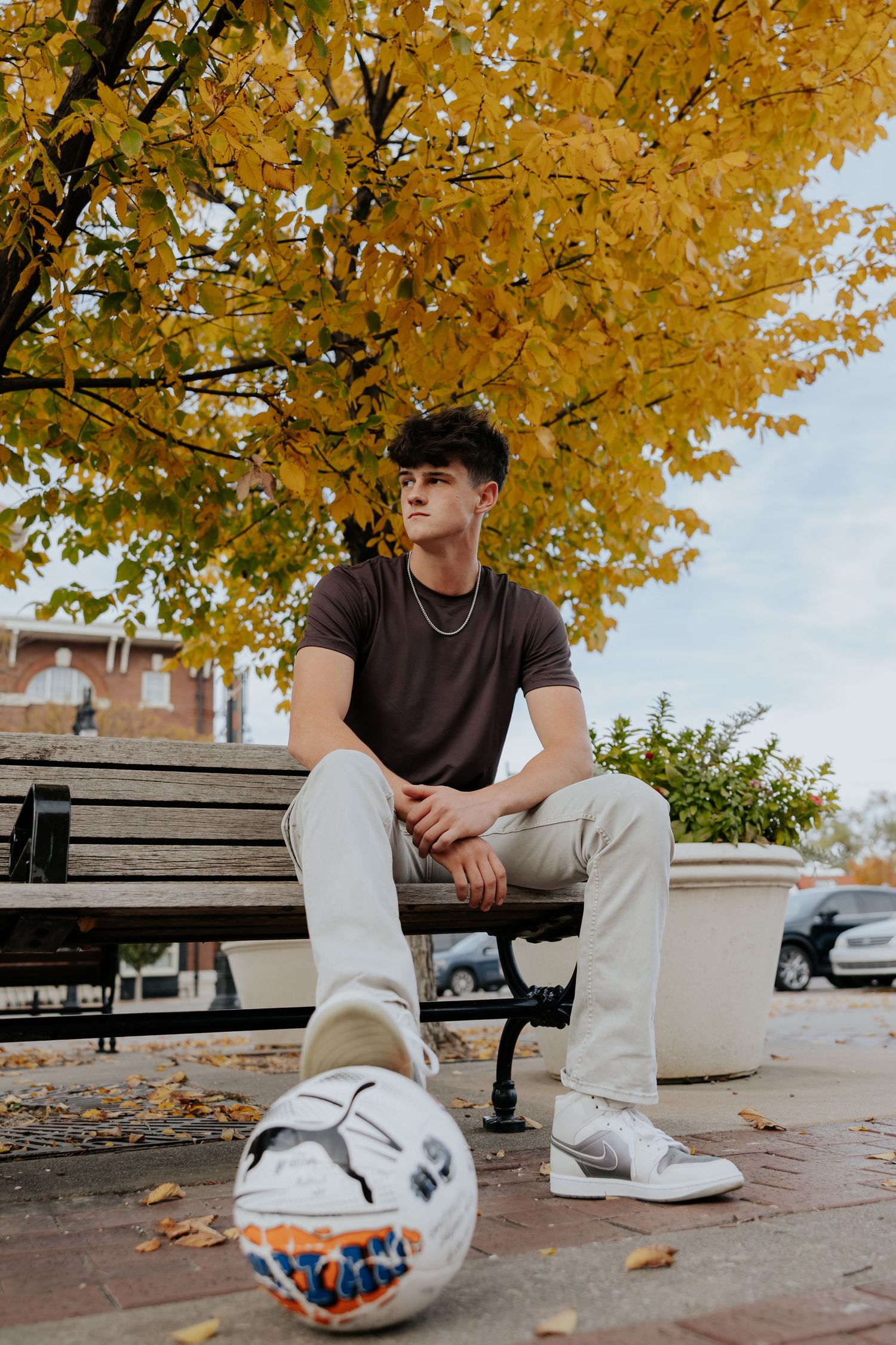 Young man in brown tee and gray pants sits on bench with soccer ball, autumn yellow leaves backdrop.