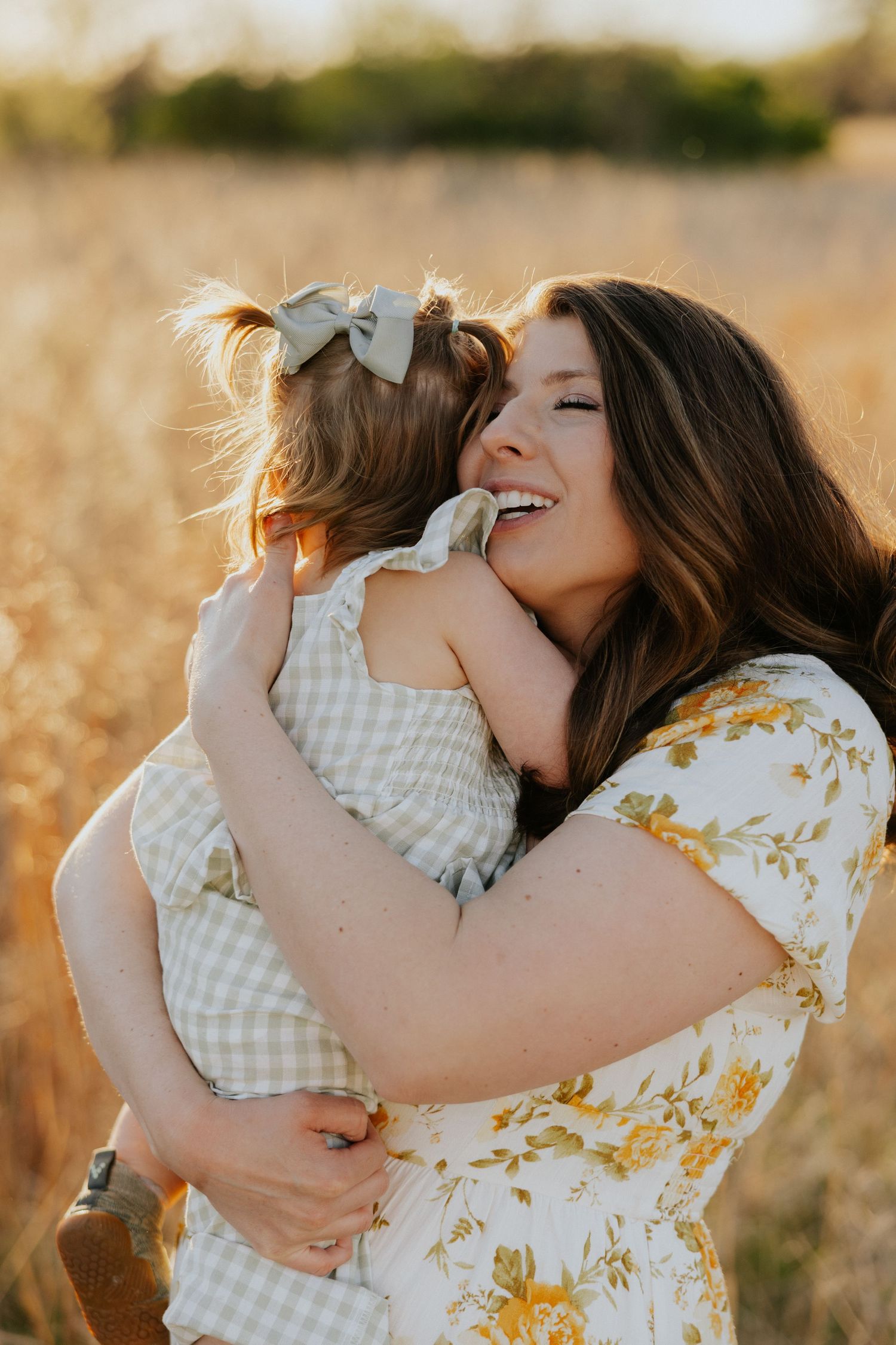 Joyful mother embraces her toddler daughter in a checkered outfit in a golden wheat field.