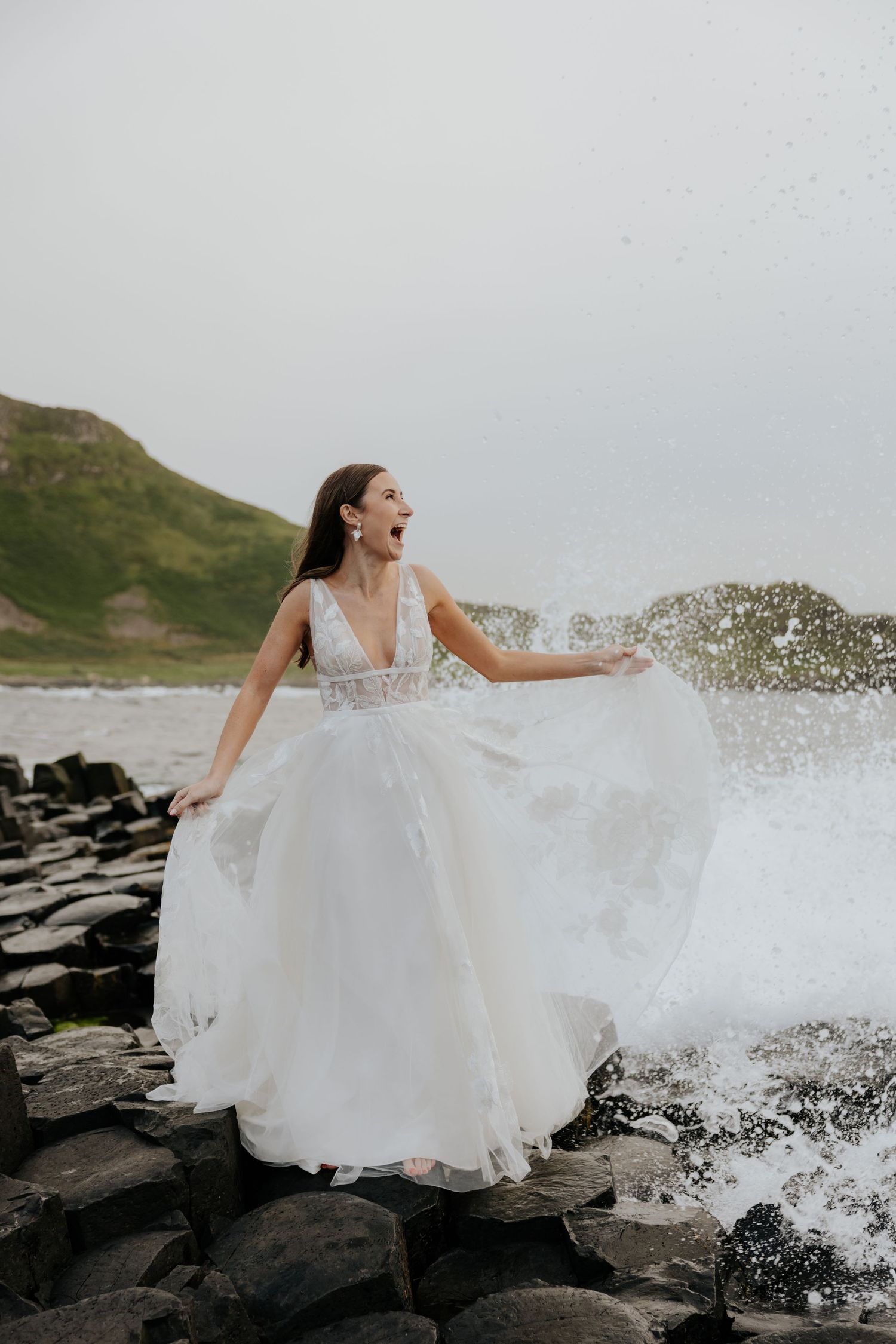 Bride in white gown laughing as ocean waves splash dramatically around her on rocky shore.