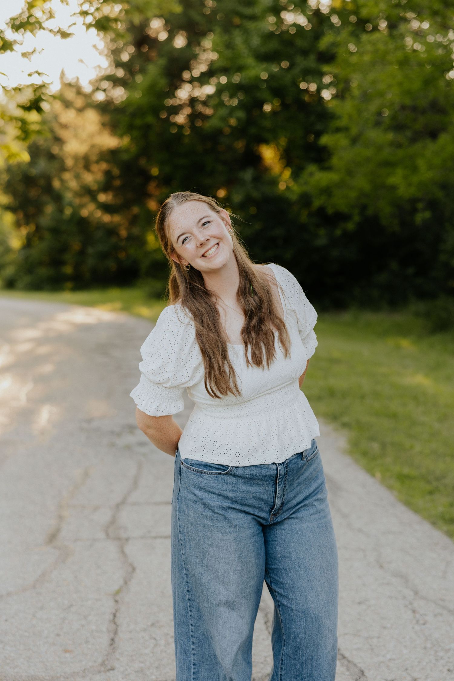 Young woman in white blouse and jeans smiling on a sunlit tree-lined path during golden hour.
