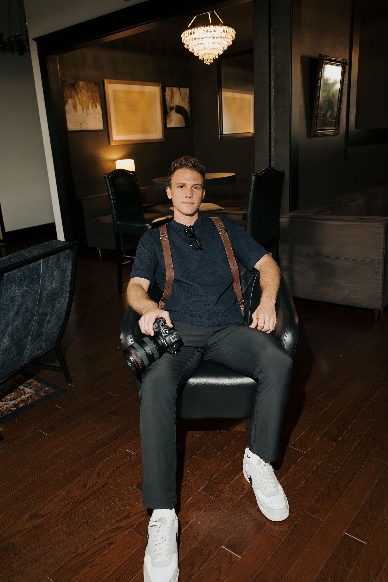 Young male photographer sitting in leather chair holding camera in elegant dark interior room.