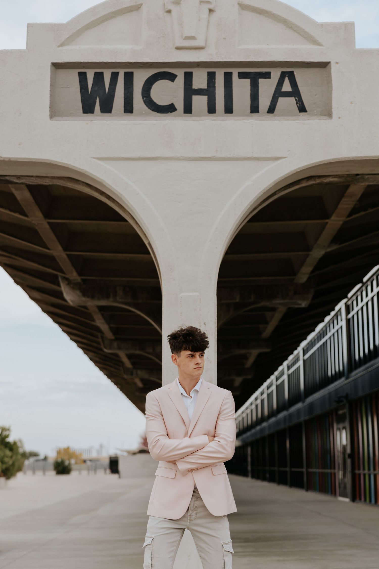 Young man in pink suit stands with arms crossed beneath Wichita train station sign outdoors.