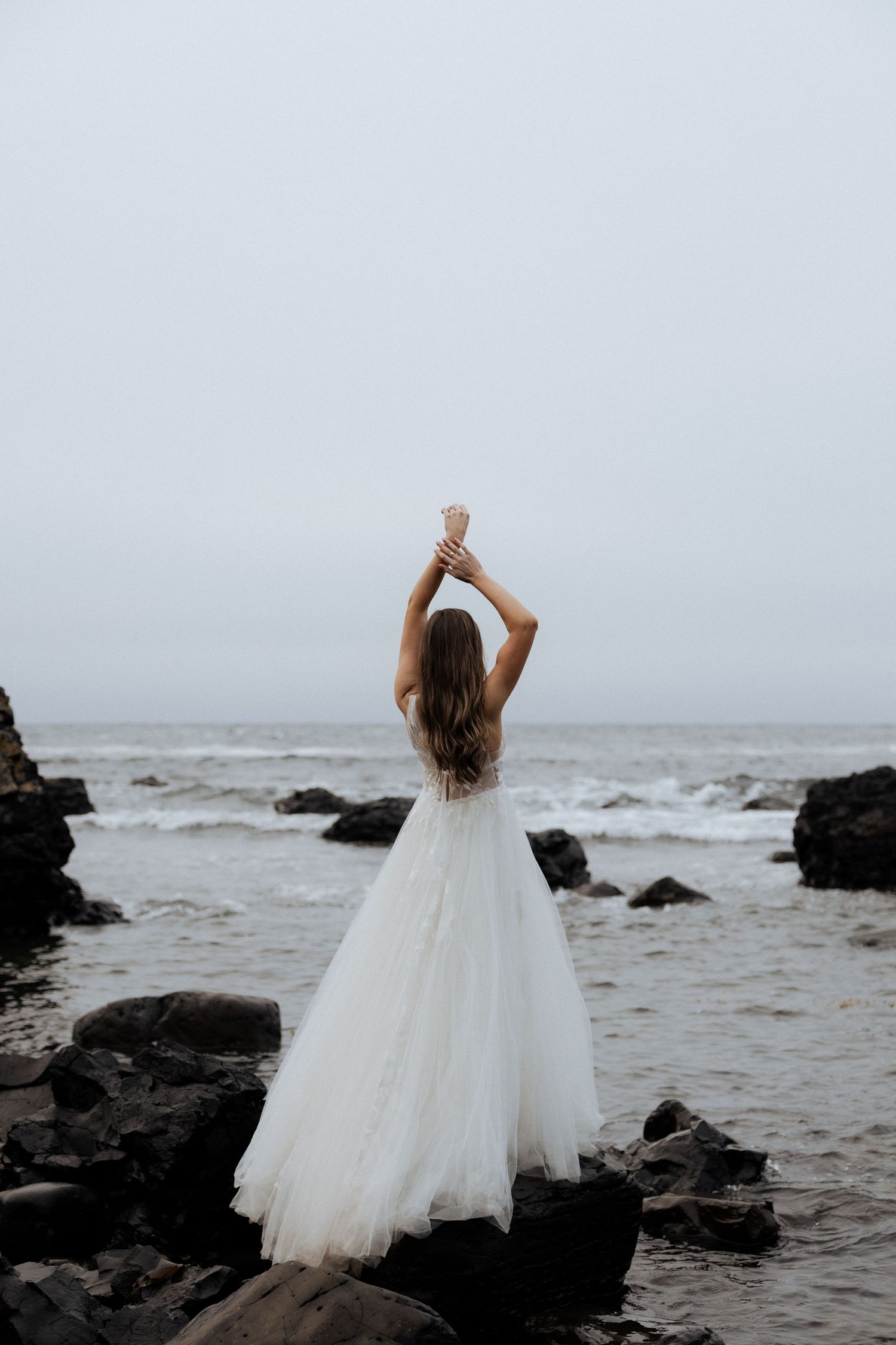 Bride in white tulle gown standing on coastal rocks with arms raised toward overcast sky.