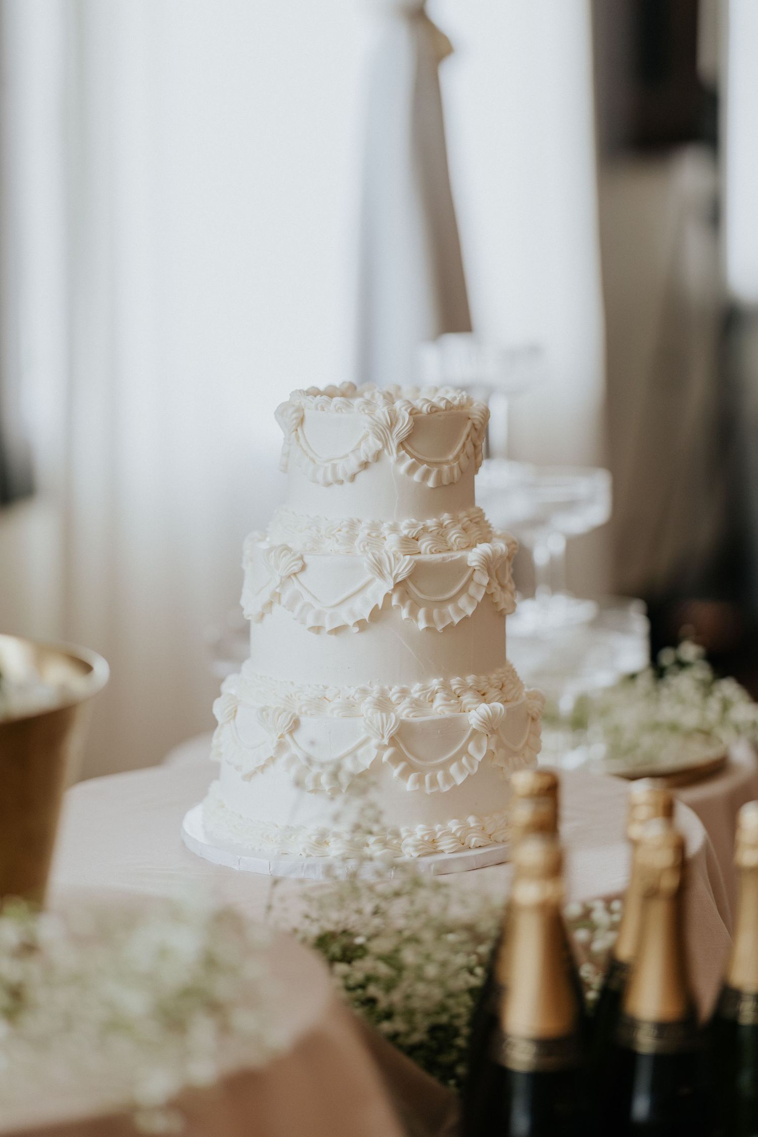 Elegant three-tier white wedding cake with ruffled details displayed on a table with champagne bottles and baby's breath flowers.