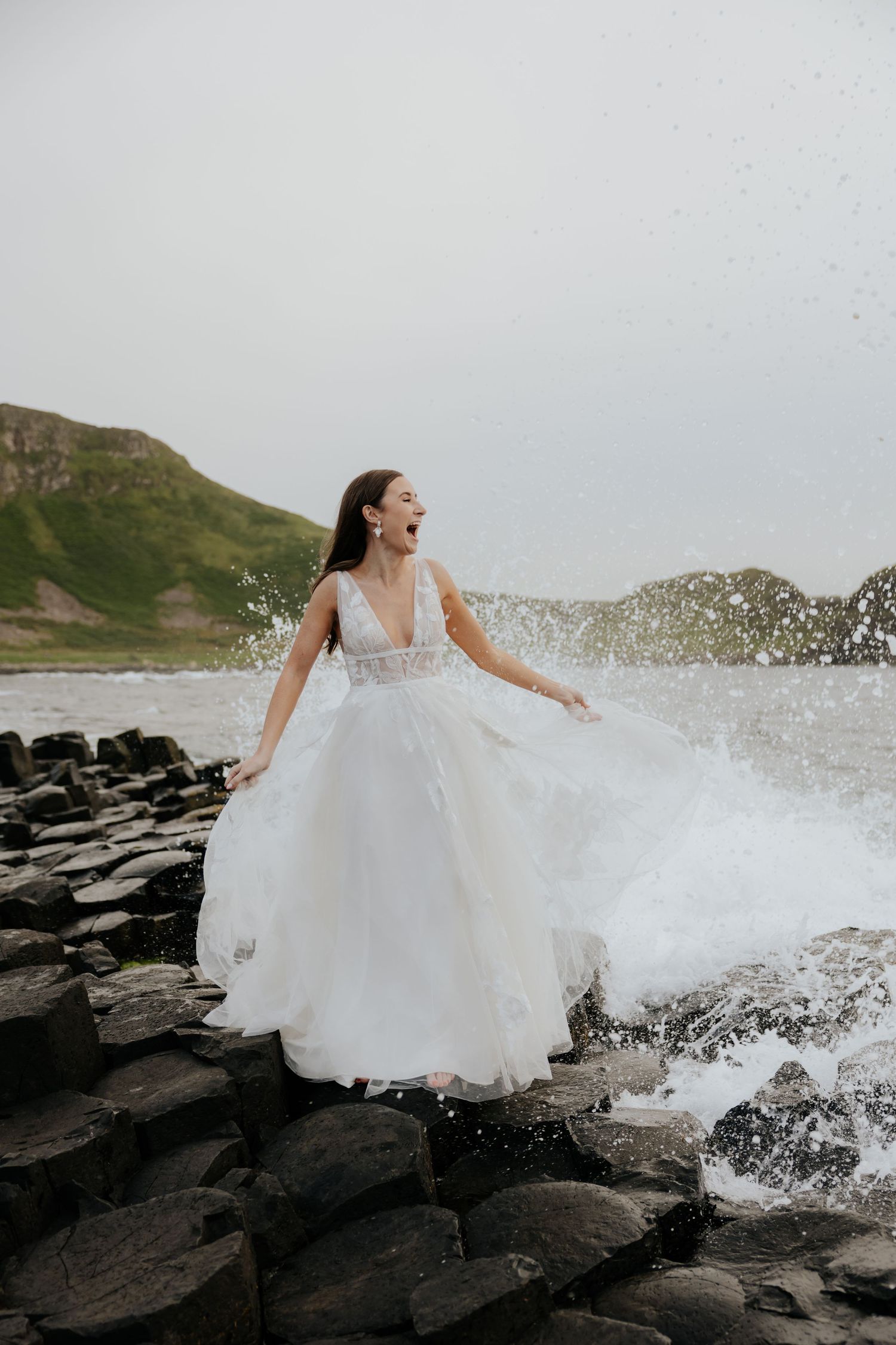 Bride in white tulle gown laughing as ocean waves splash around her on rocky coastal basalt columns.