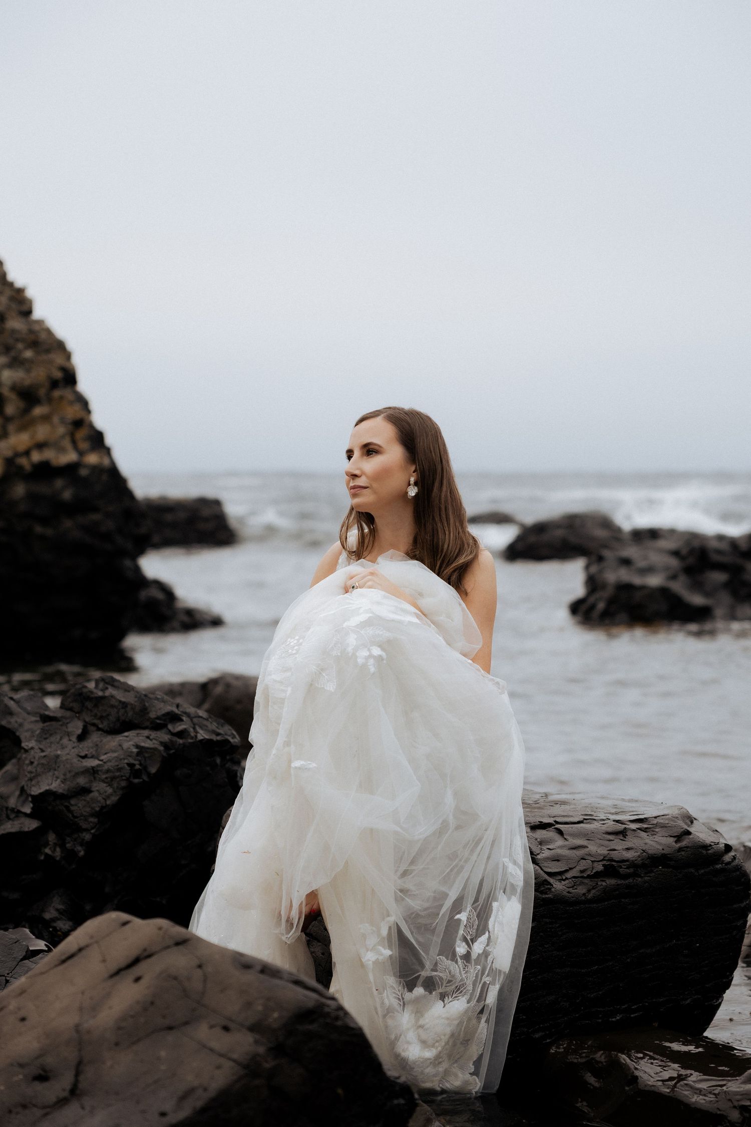 Bride in strapless white tulle gown standing among dark rocks with moody ocean scenery.