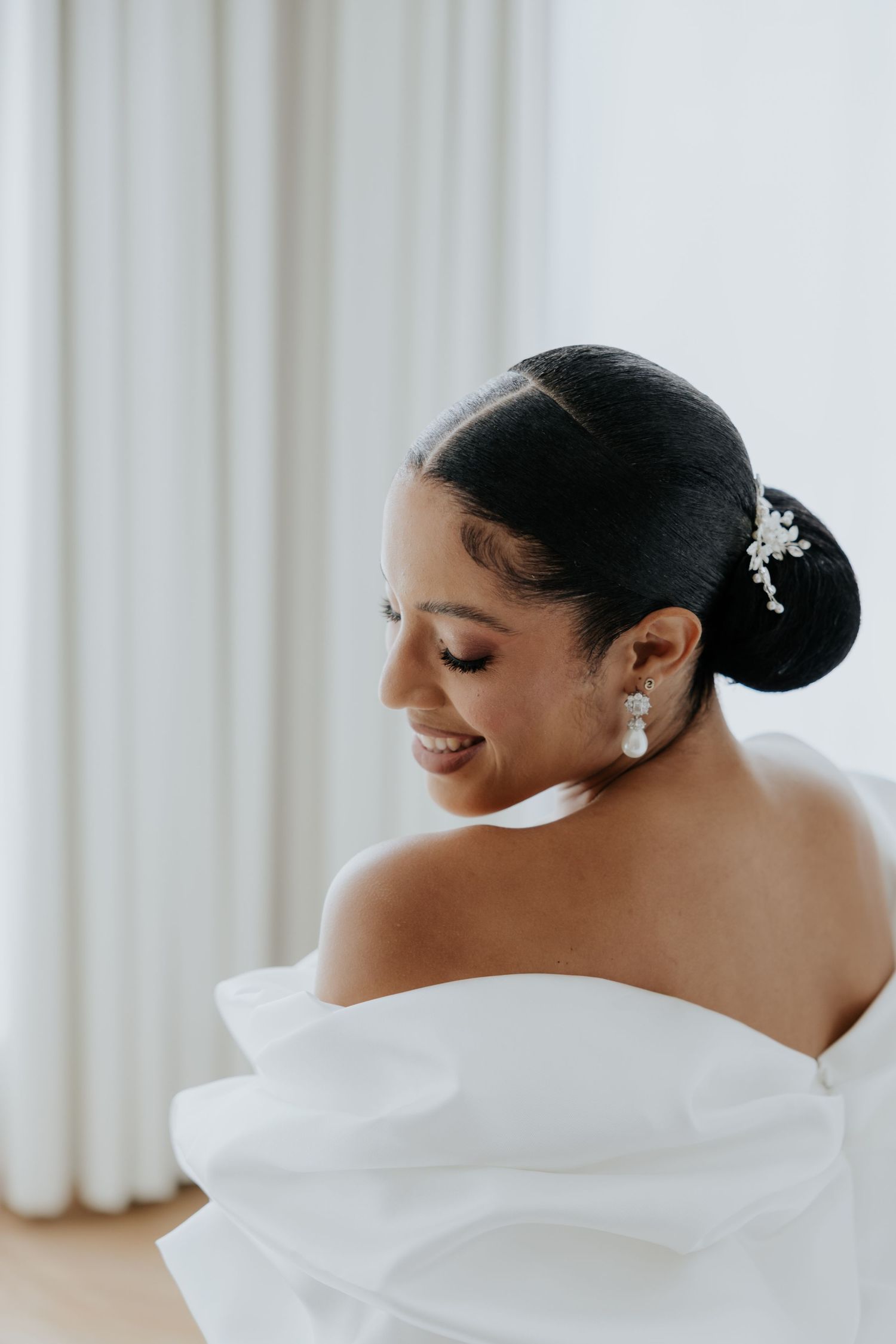 Bride with elegant updo and pearl earrings smiles in white off-shoulder wedding gown.