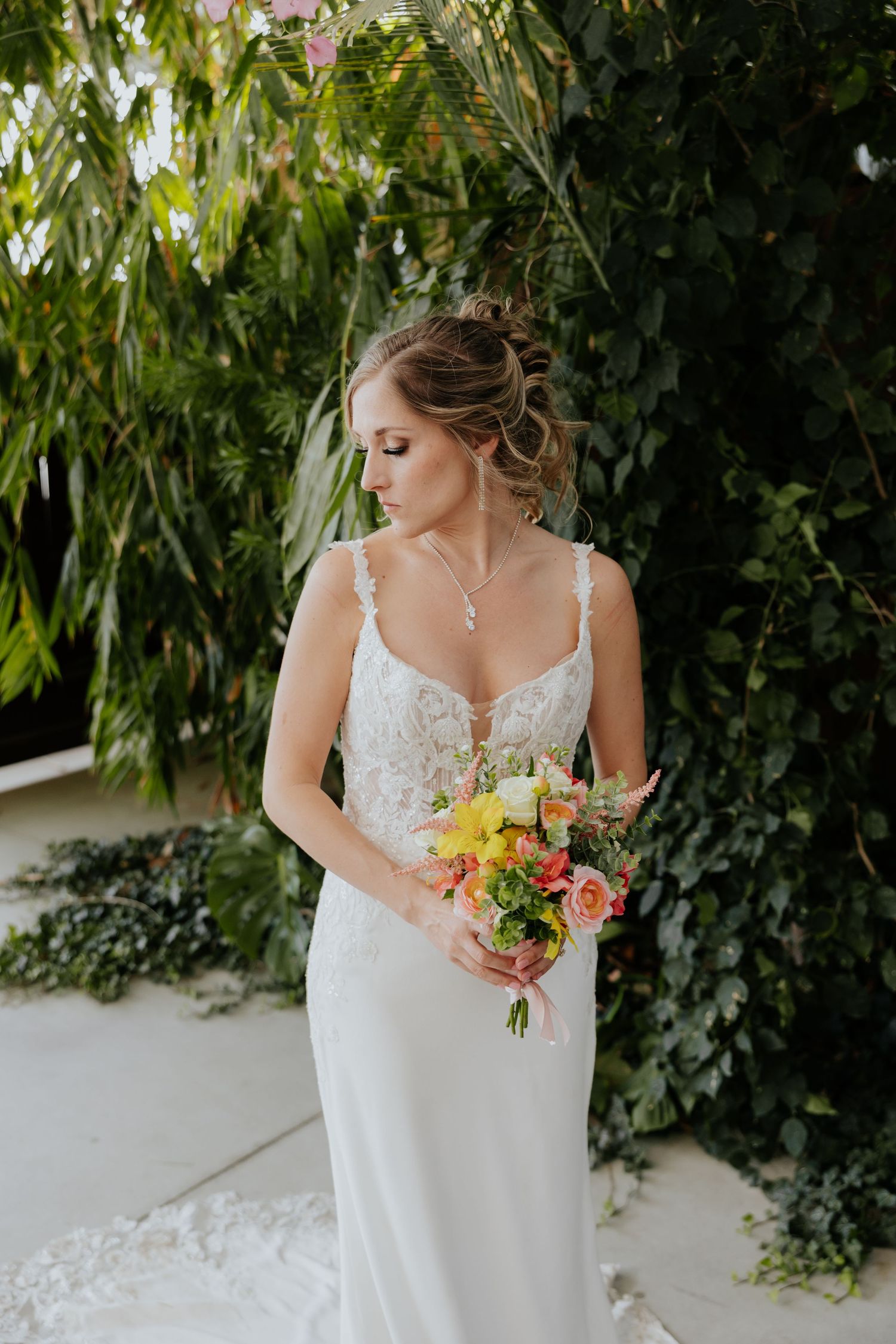Bride in lace wedding gown holding colorful bouquet of roses standing before lush green foliage.