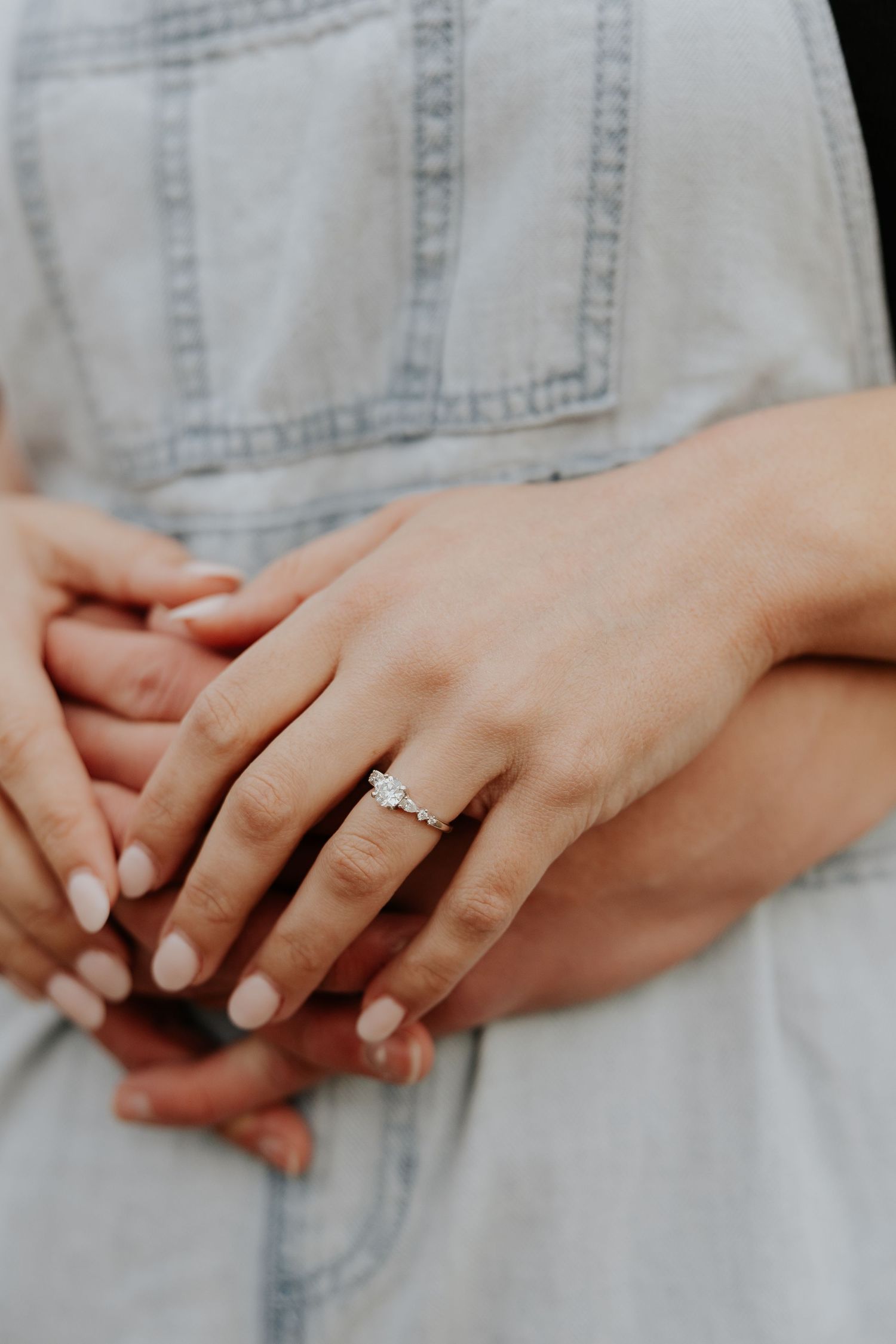 Close-up of an elegant diamond engagement ring on woman's hand wearing light denim overalls outdoors.